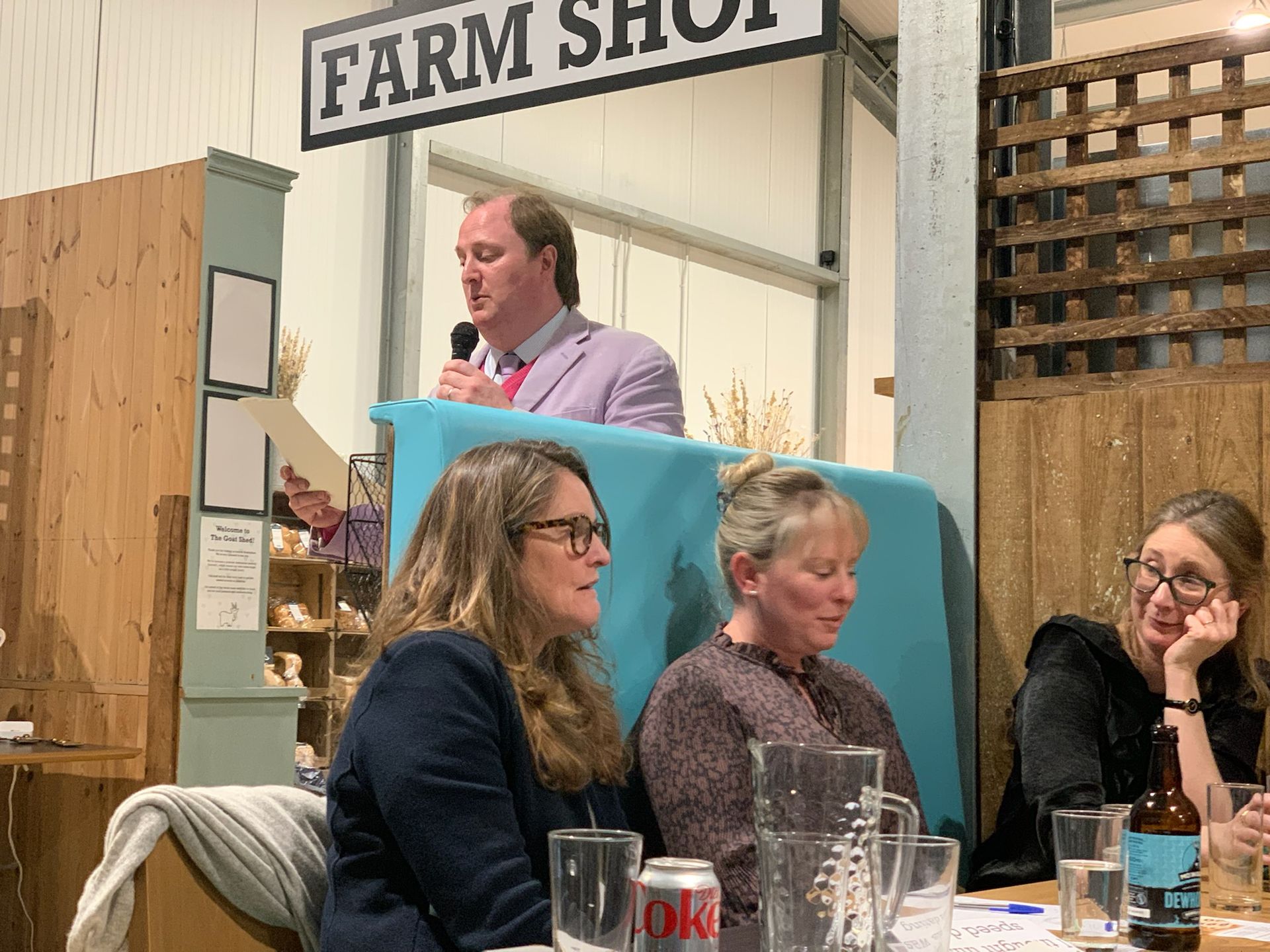 Auctioneer speaking above three seated women at a Farm Shop, holding microphone.