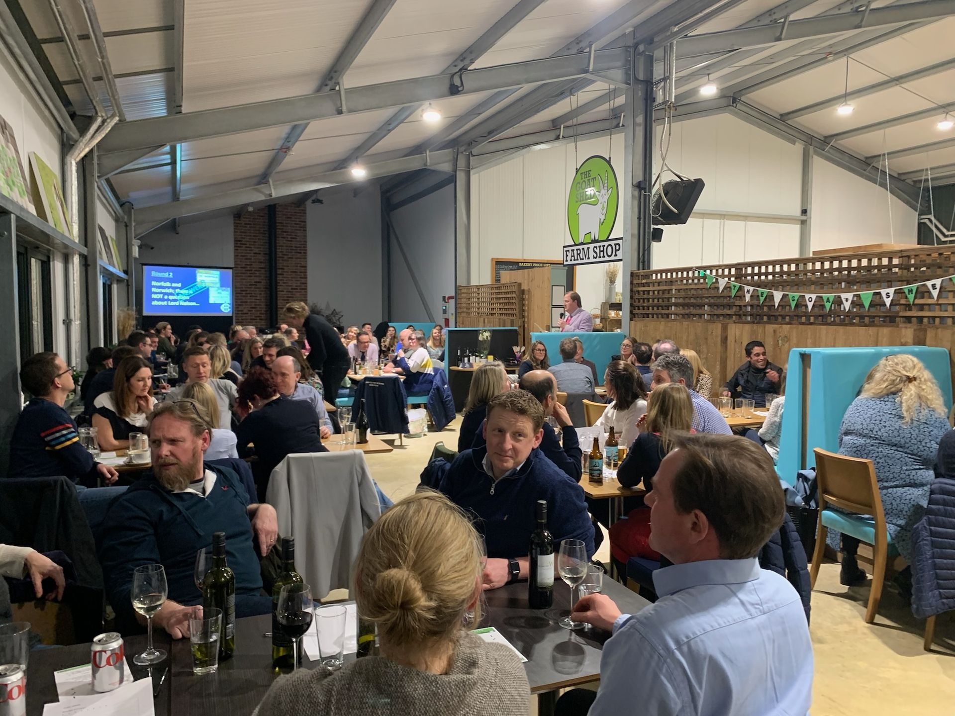 Large gathering at tables in a brightly lit room. People are eating and socializing, with a screen and a logo visible.