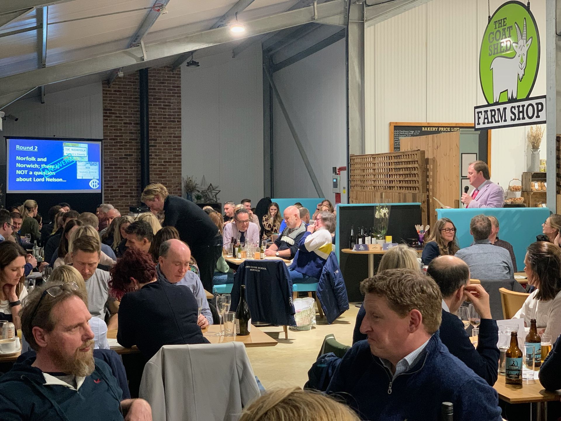 People gathered in a large room, some seated, with a man speaking at a podium. A TV screen shows text.