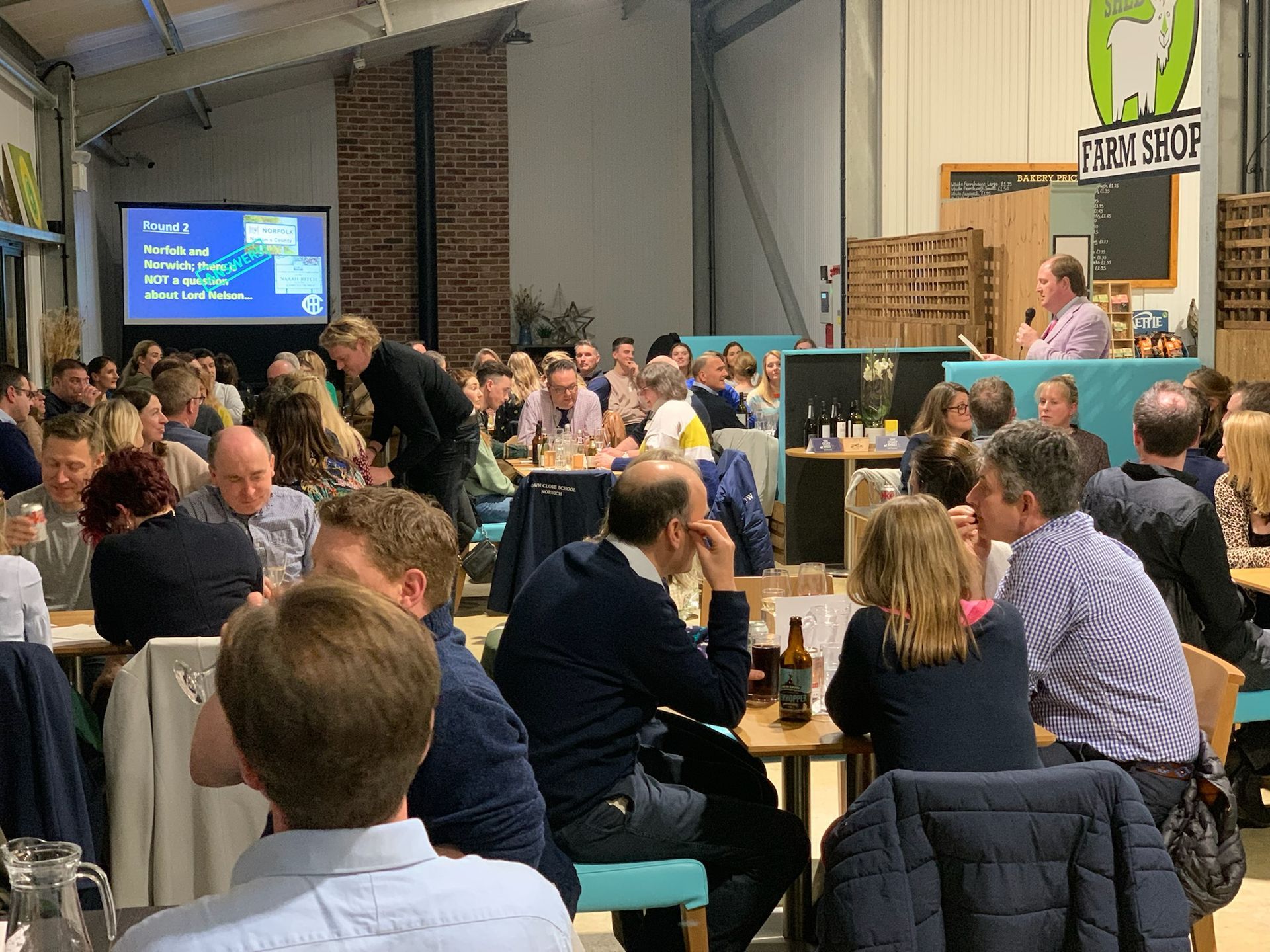 Large gathering in a warehouse-like space; people seated at tables, a speaker at the front, and a screen with text.