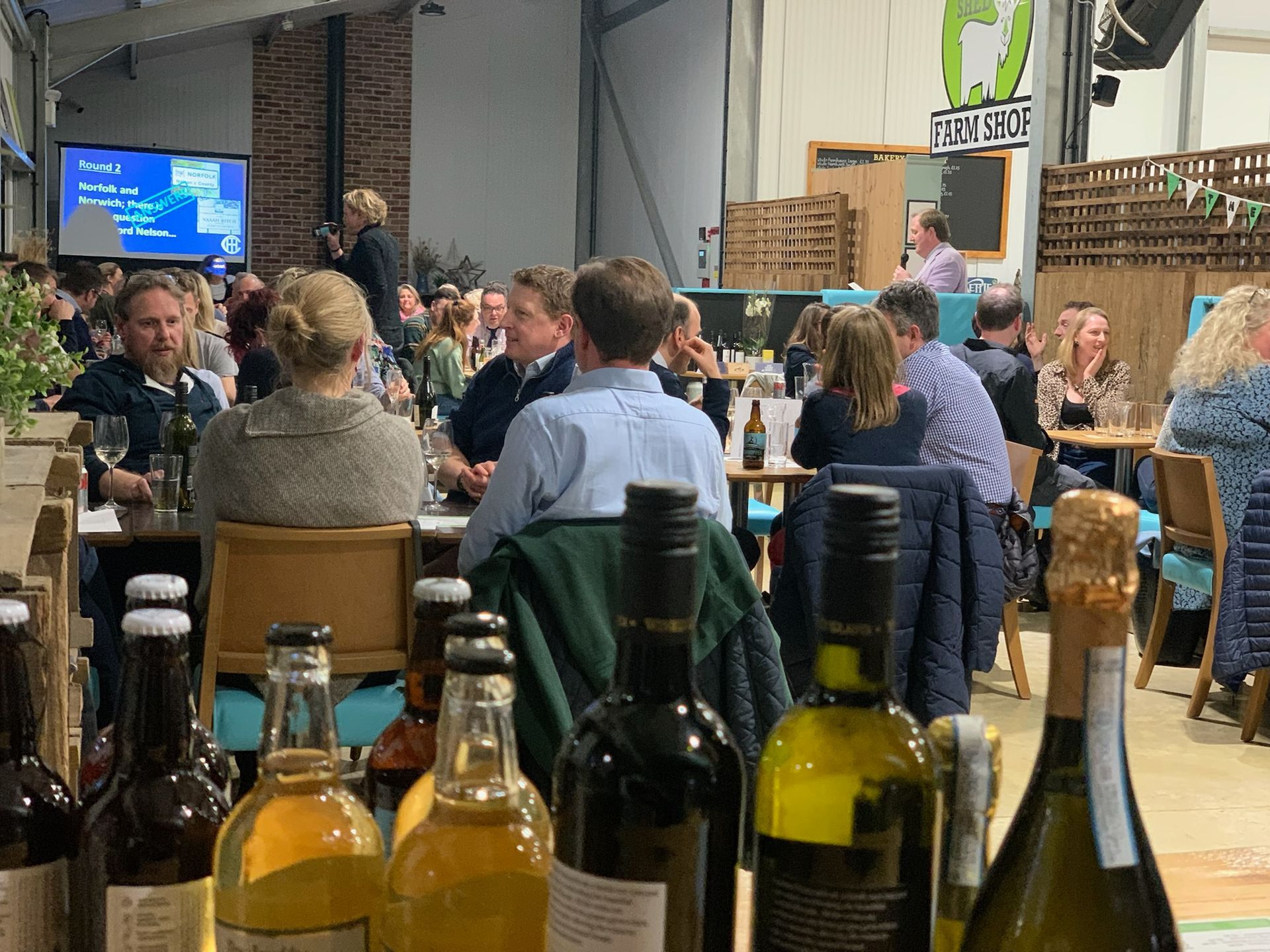 People at tables in a large venue, bottles of drinks in front, speaker on screen.