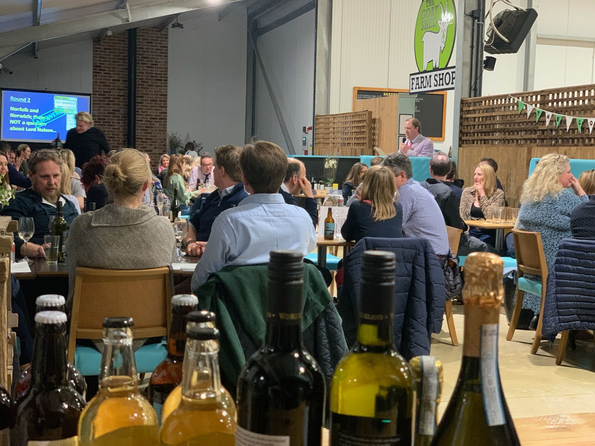 People at tables in a brewery listen to a speaker, bottles in the foreground.