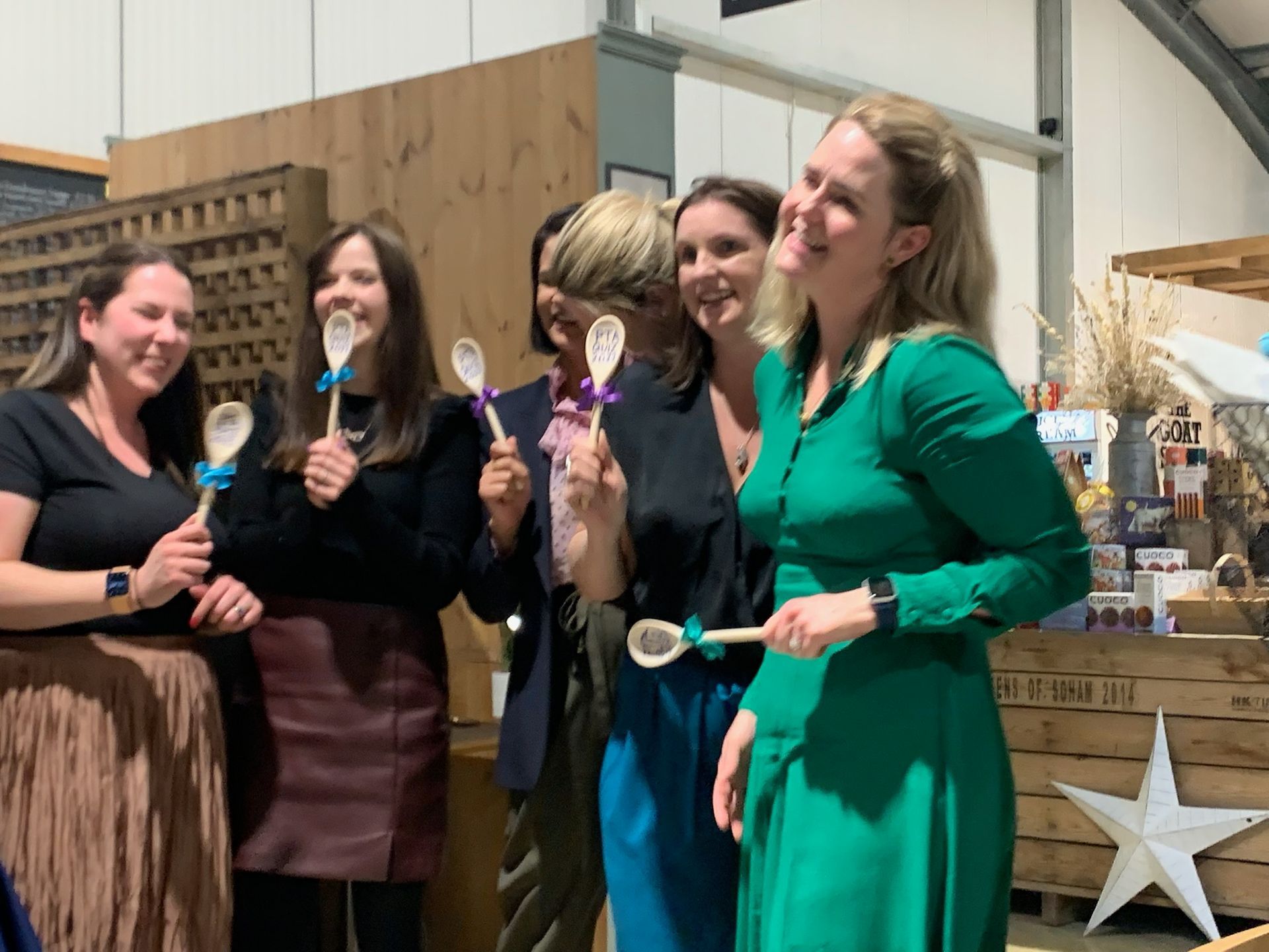 Six women holding wooden spoons, smiling. Indoor market setting, some wearing dresses and blazers.