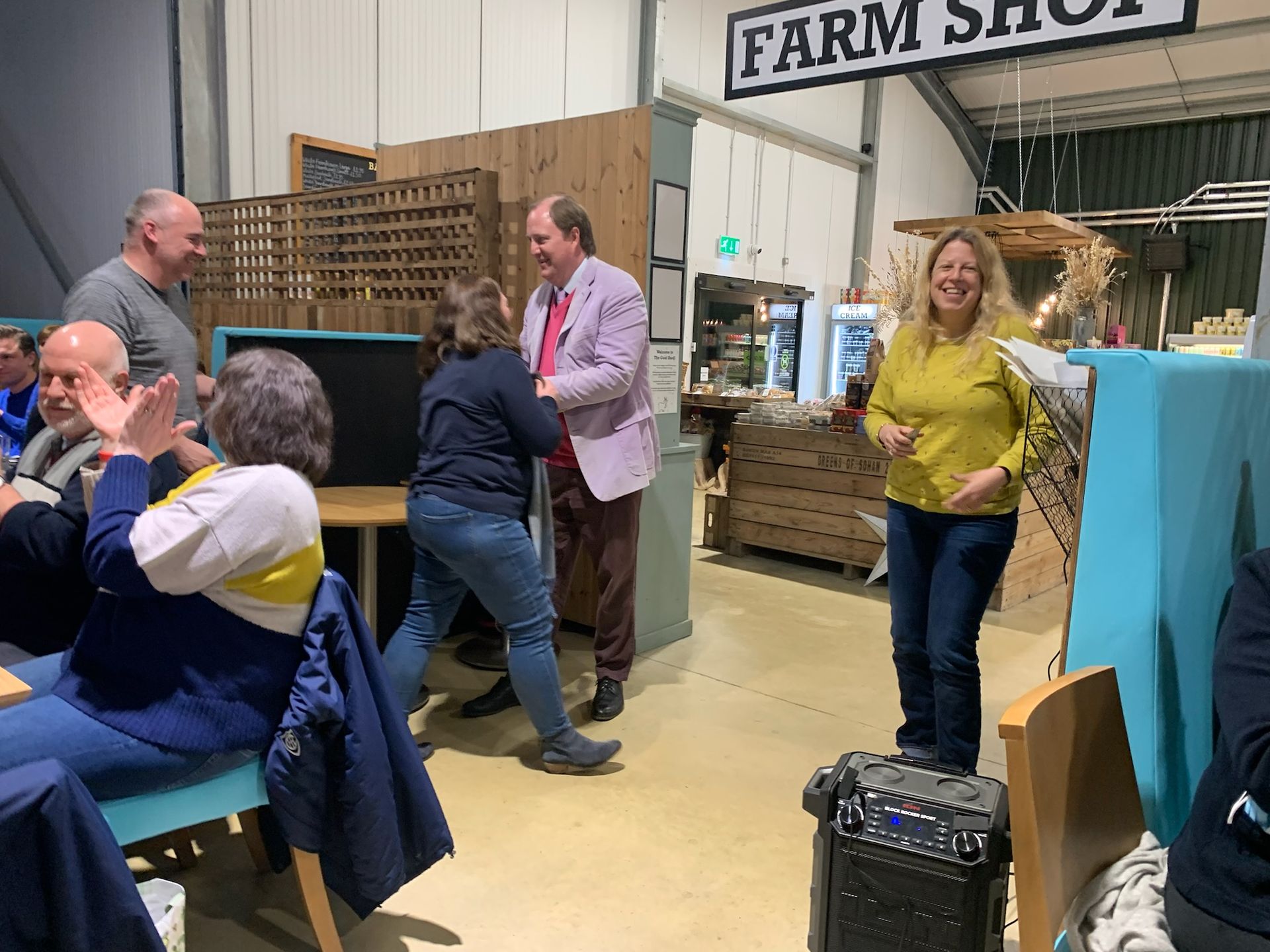 People in a farm shop, some clapping. A woman is dancing while a man smiles.