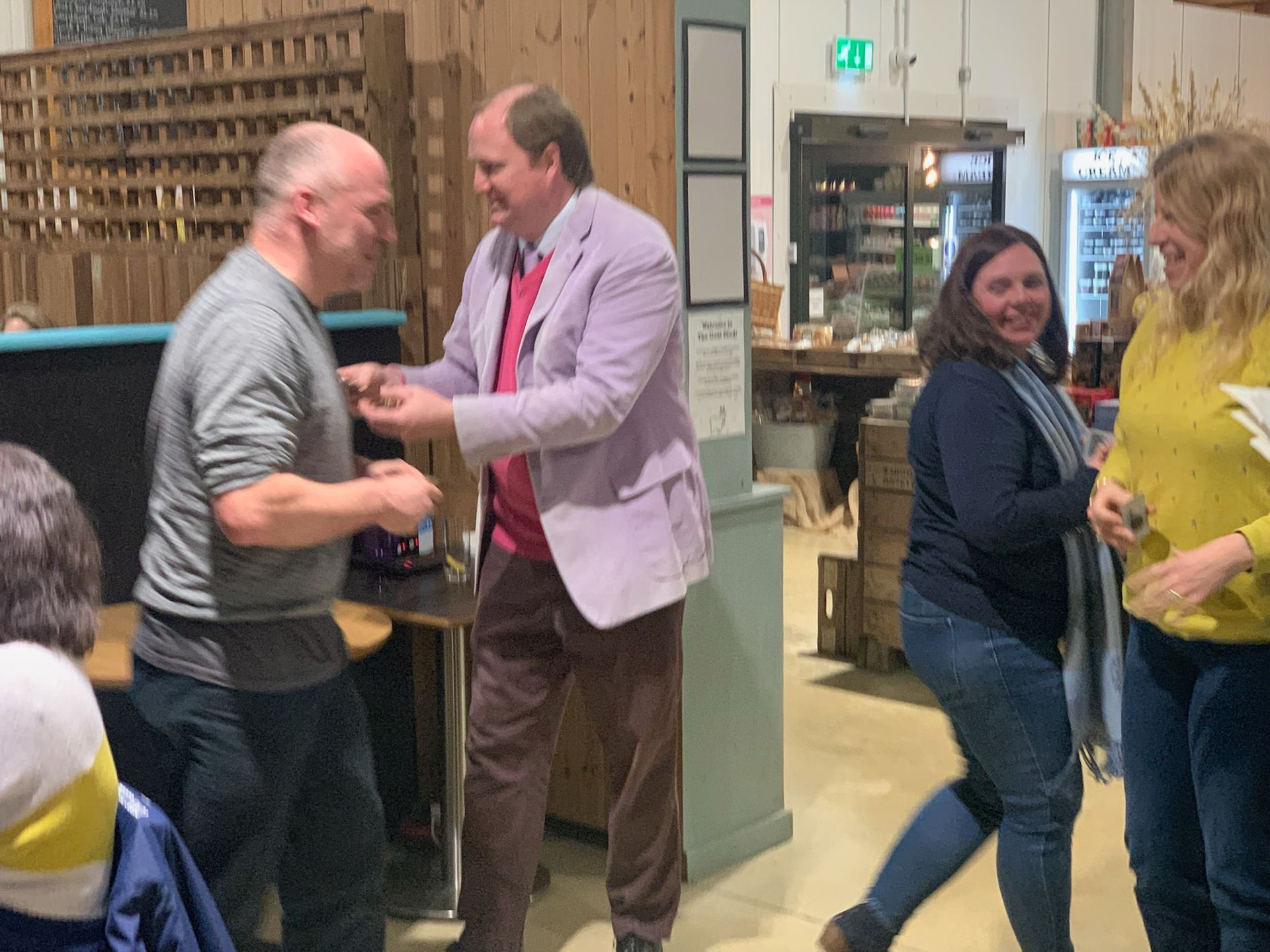 Man in pink blazer shaking hands with another man; two women smile nearby in a casual setting.