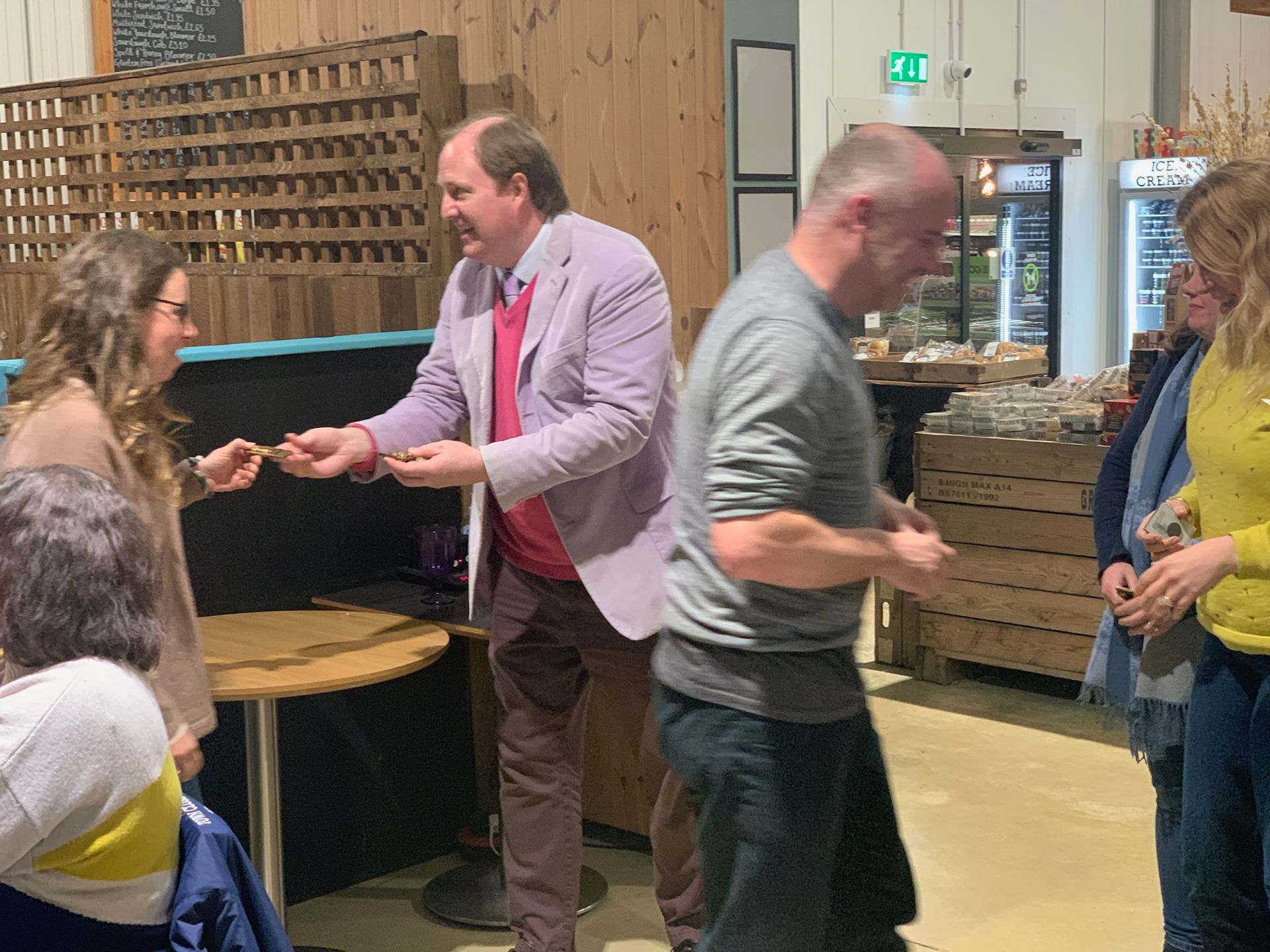 Man in lavender jacket handing something to woman, others watch in a market-like setting.