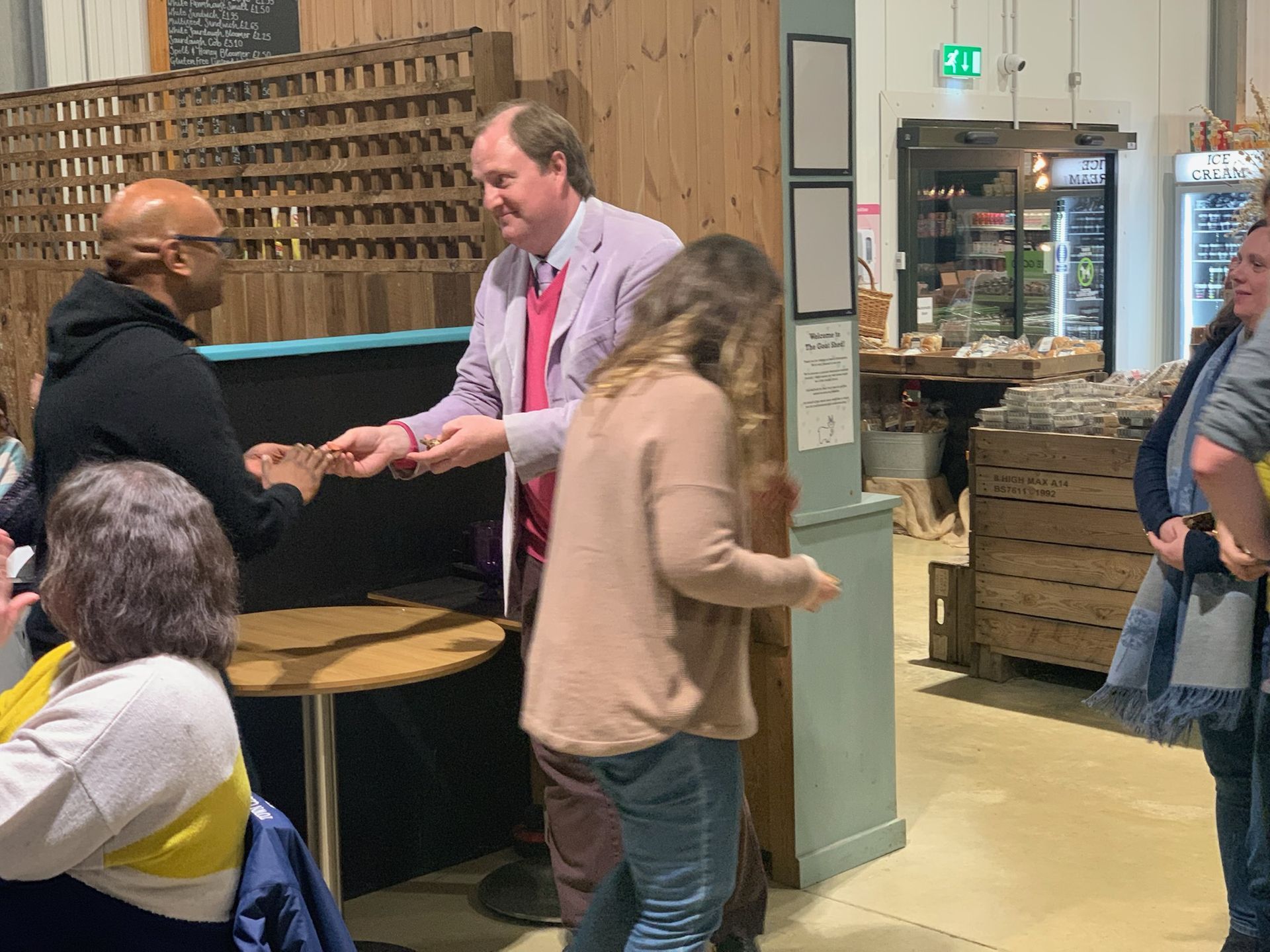 Man in pink jacket hands something to another man; people watch in a cafe.