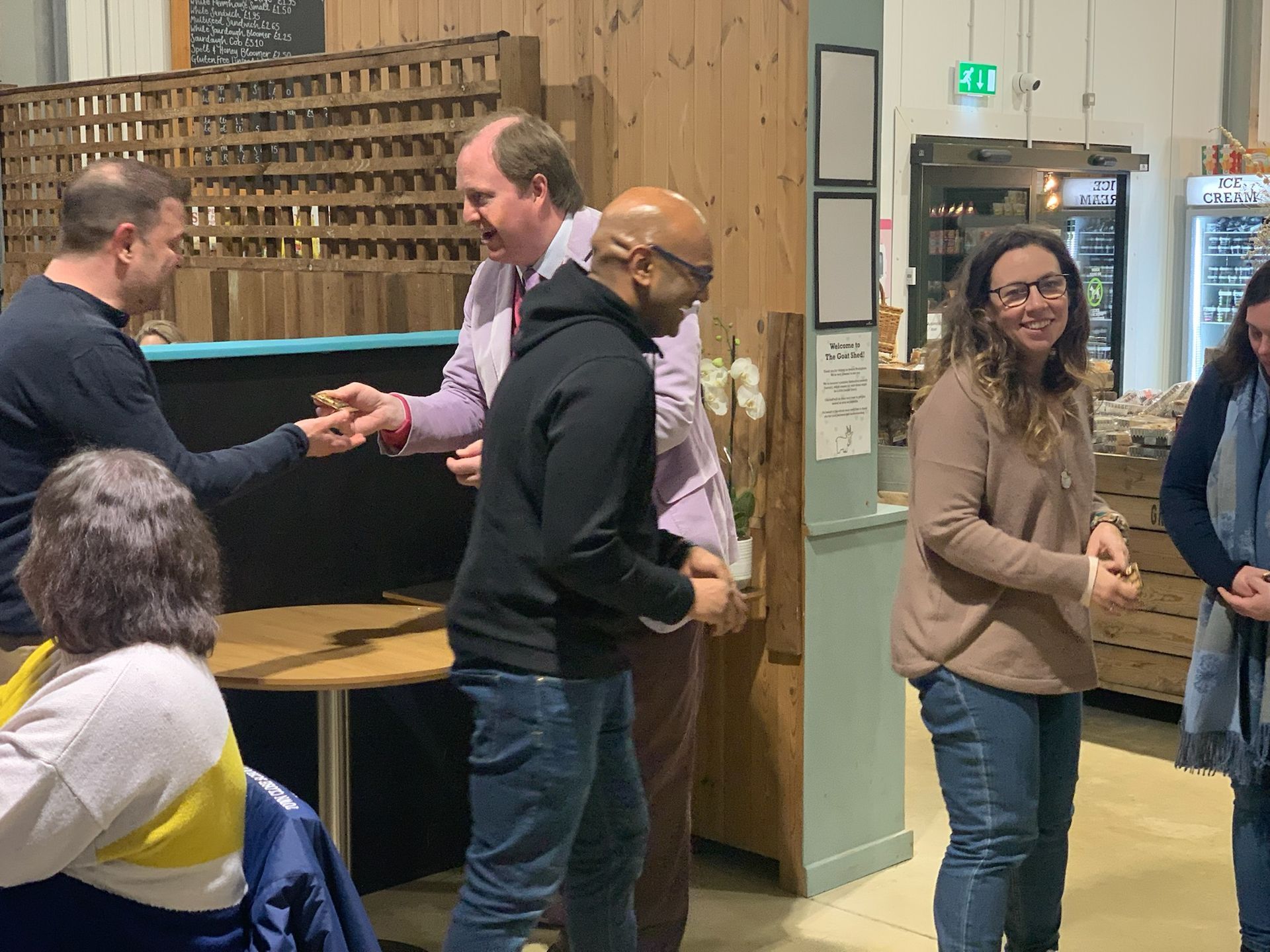 People in a store: two men shake hands, others watch, smiling. Store has wood and white accents.