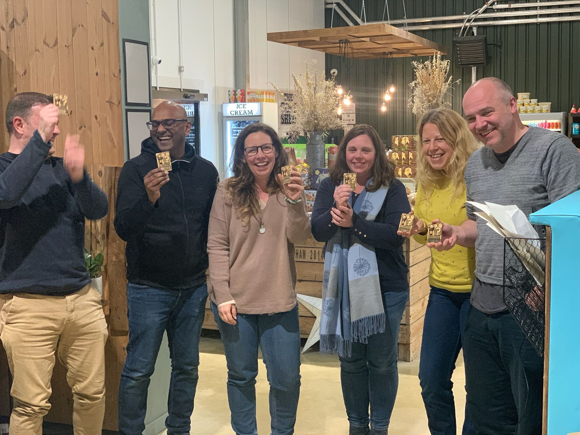 Six people smiling and holding small wooden trophies indoors, celebrating an event.