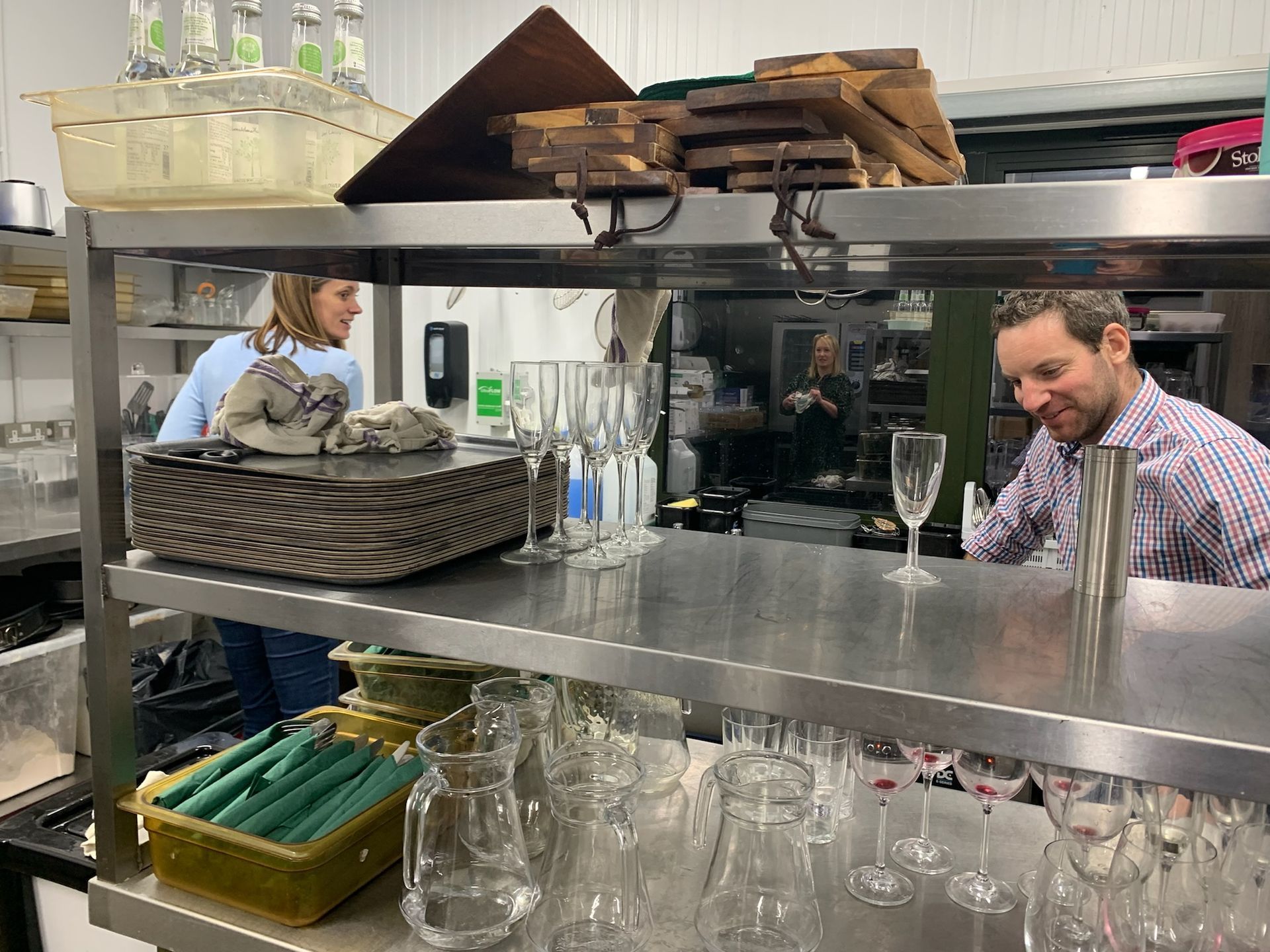 Restaurant kitchen with staff working, stainless steel shelves, glasses, and dishware.