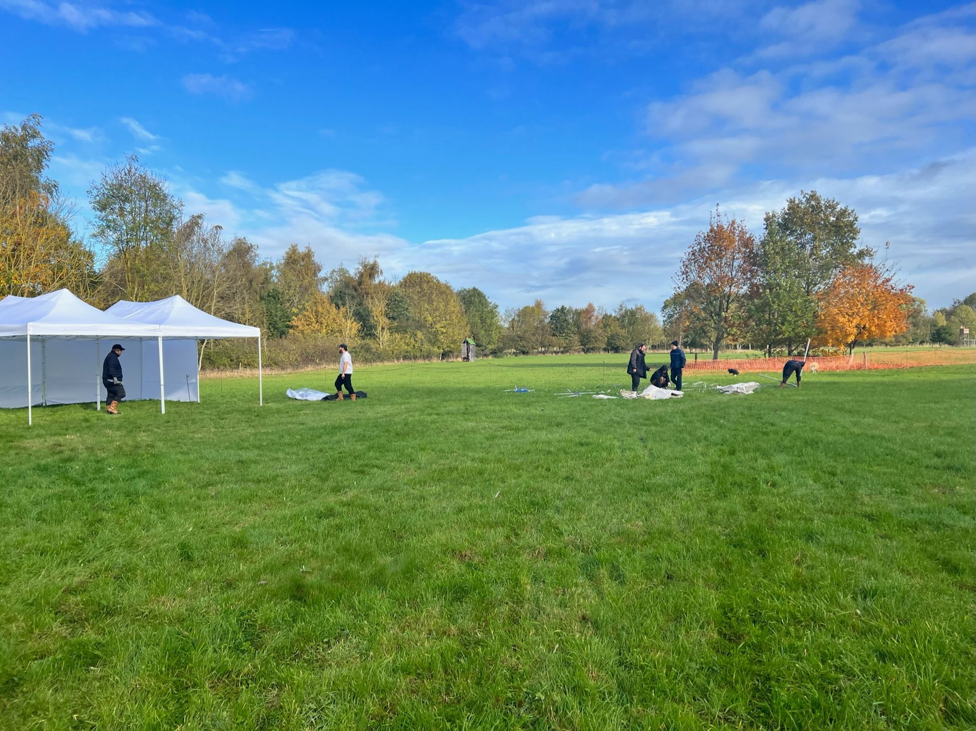 Green park scene; white tent on left, people setting up near trees under blue sky.