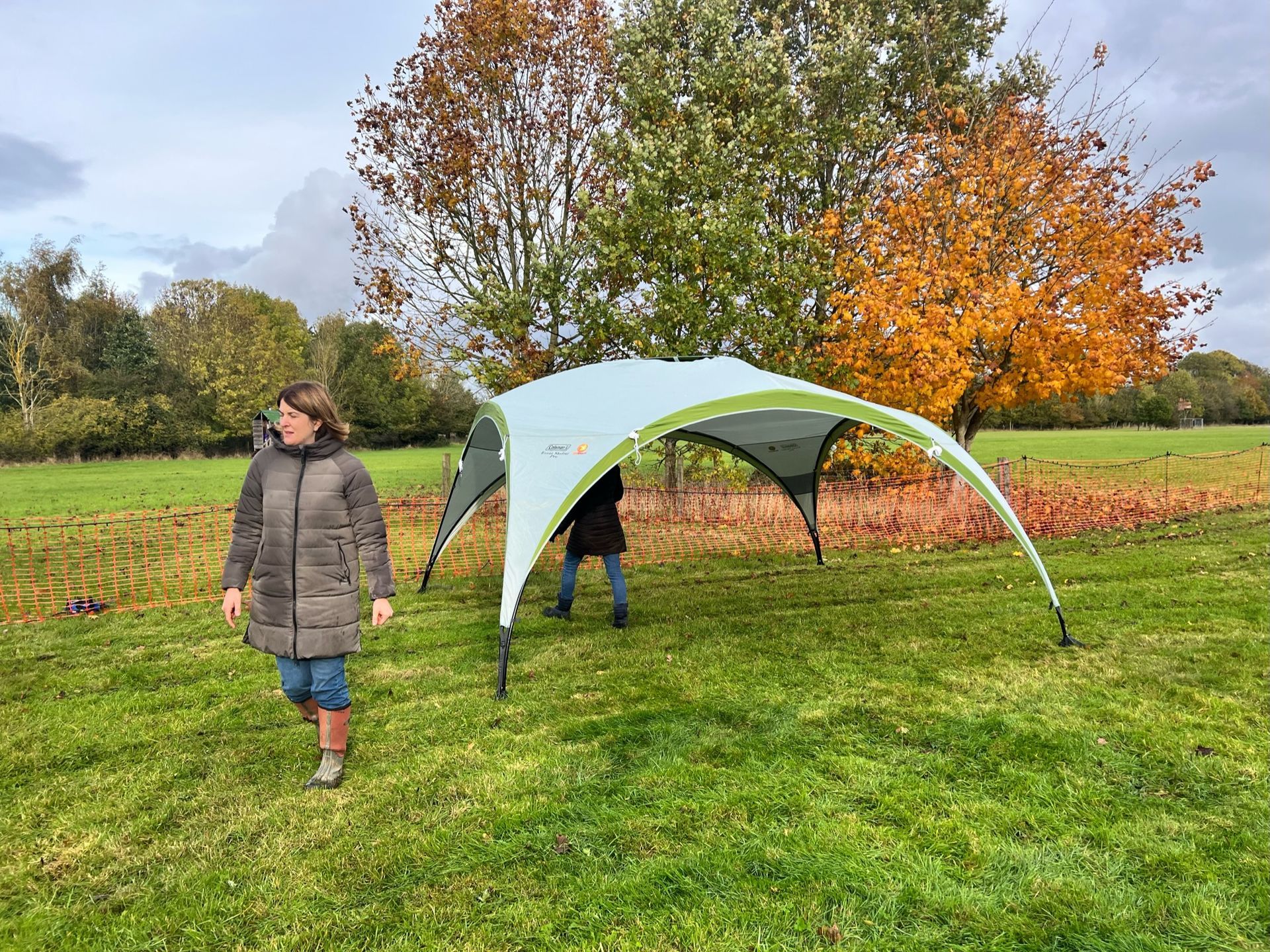 Woman walks past a light-blue and green pop-up tent on a grassy field, fall trees in background.