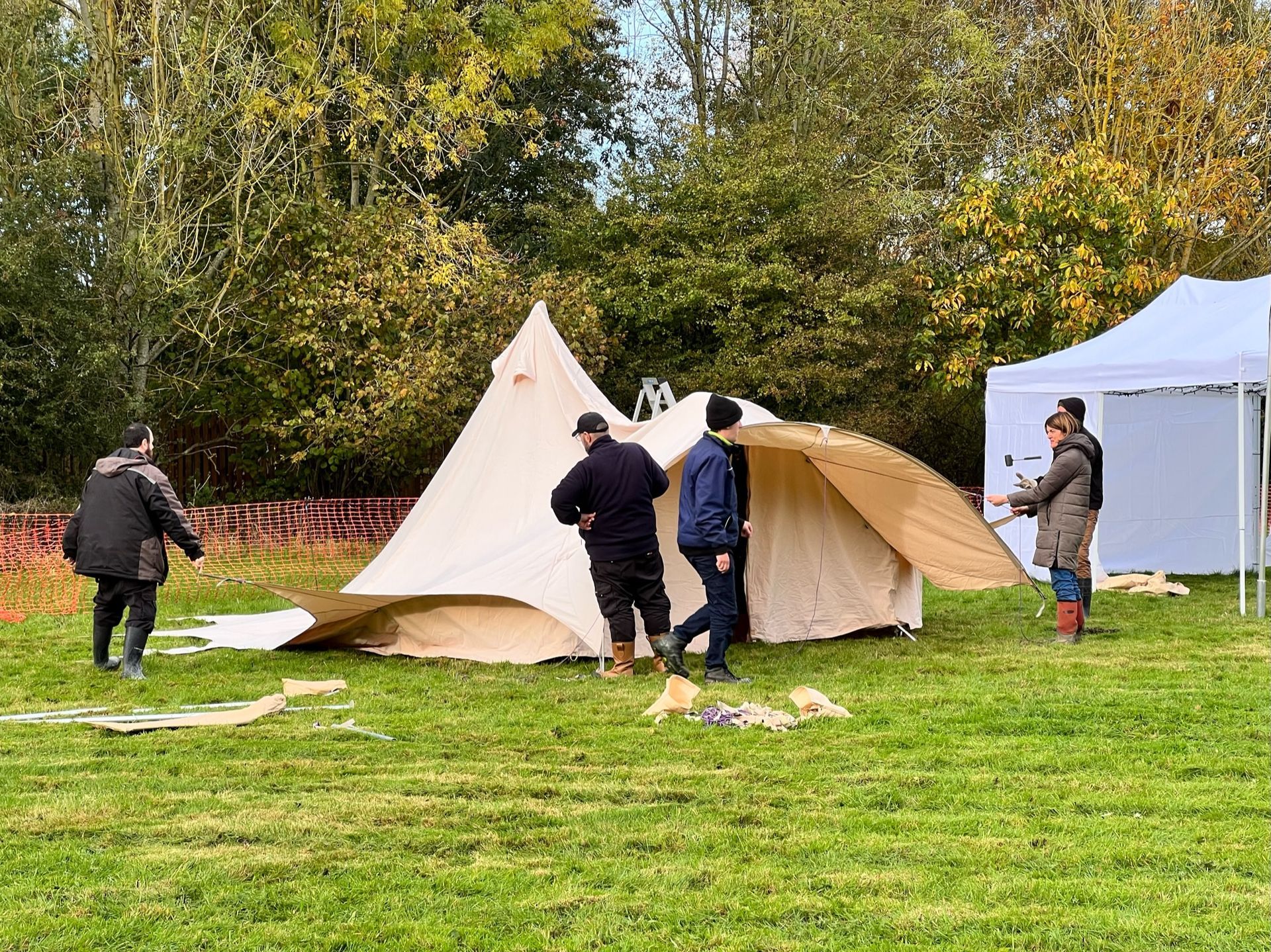 People assembling a large tan canvas tent in a grassy field with trees in the background.