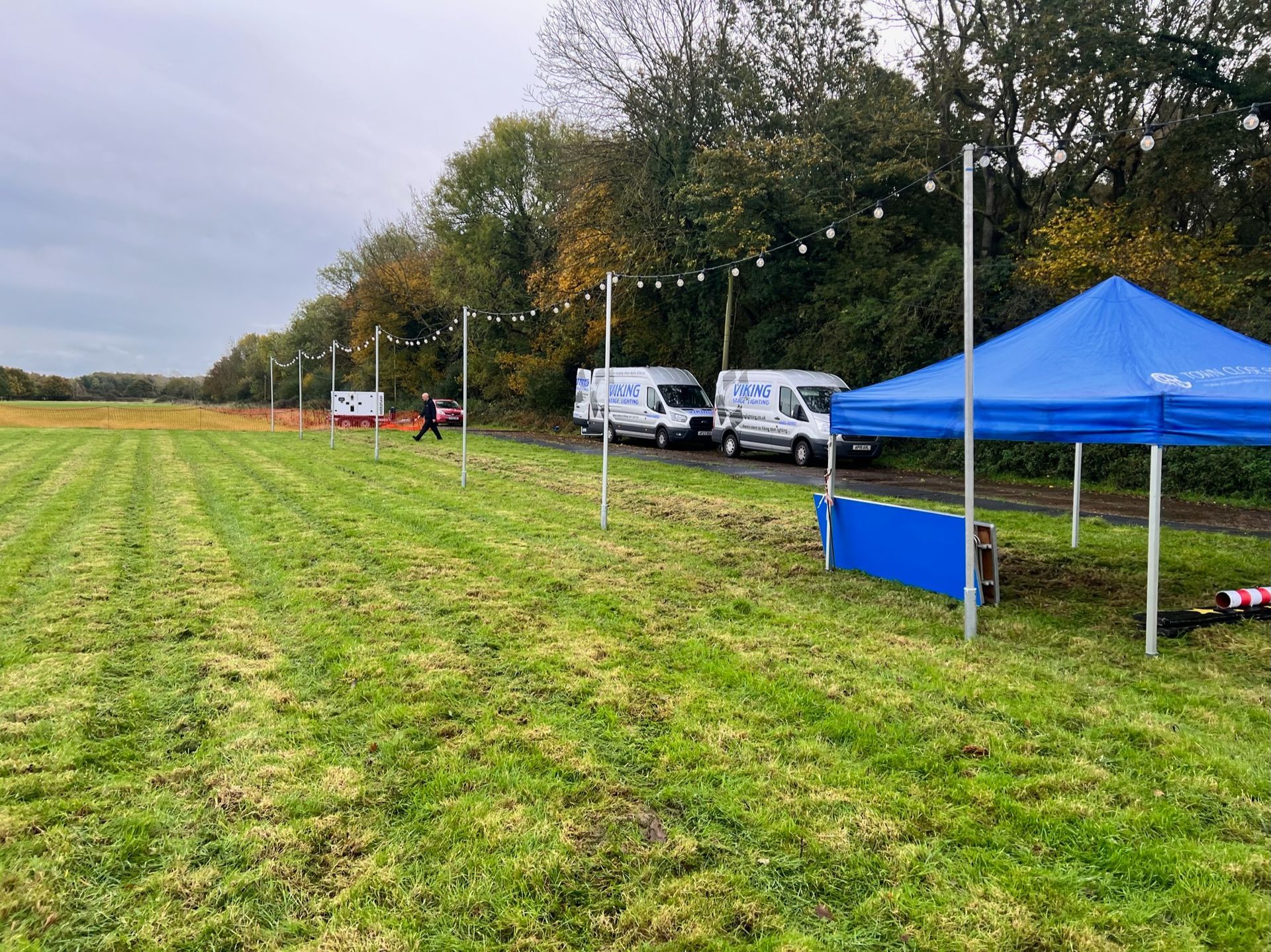 Grassy field with parked vans, a blue tent, string lights, and a distant figure under overcast skies.