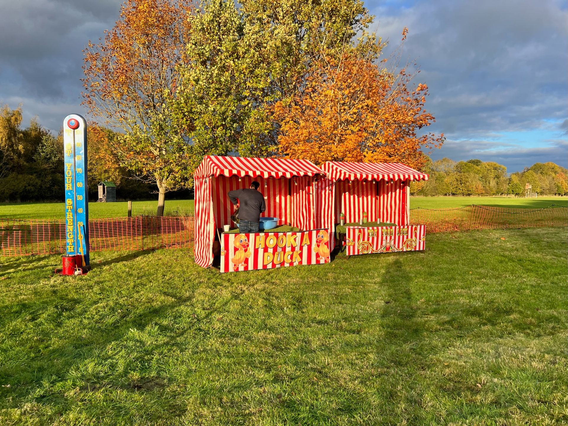 Person setting up a red and white striped stall in a park with a tall temperature gauge; autumn trees in background.
