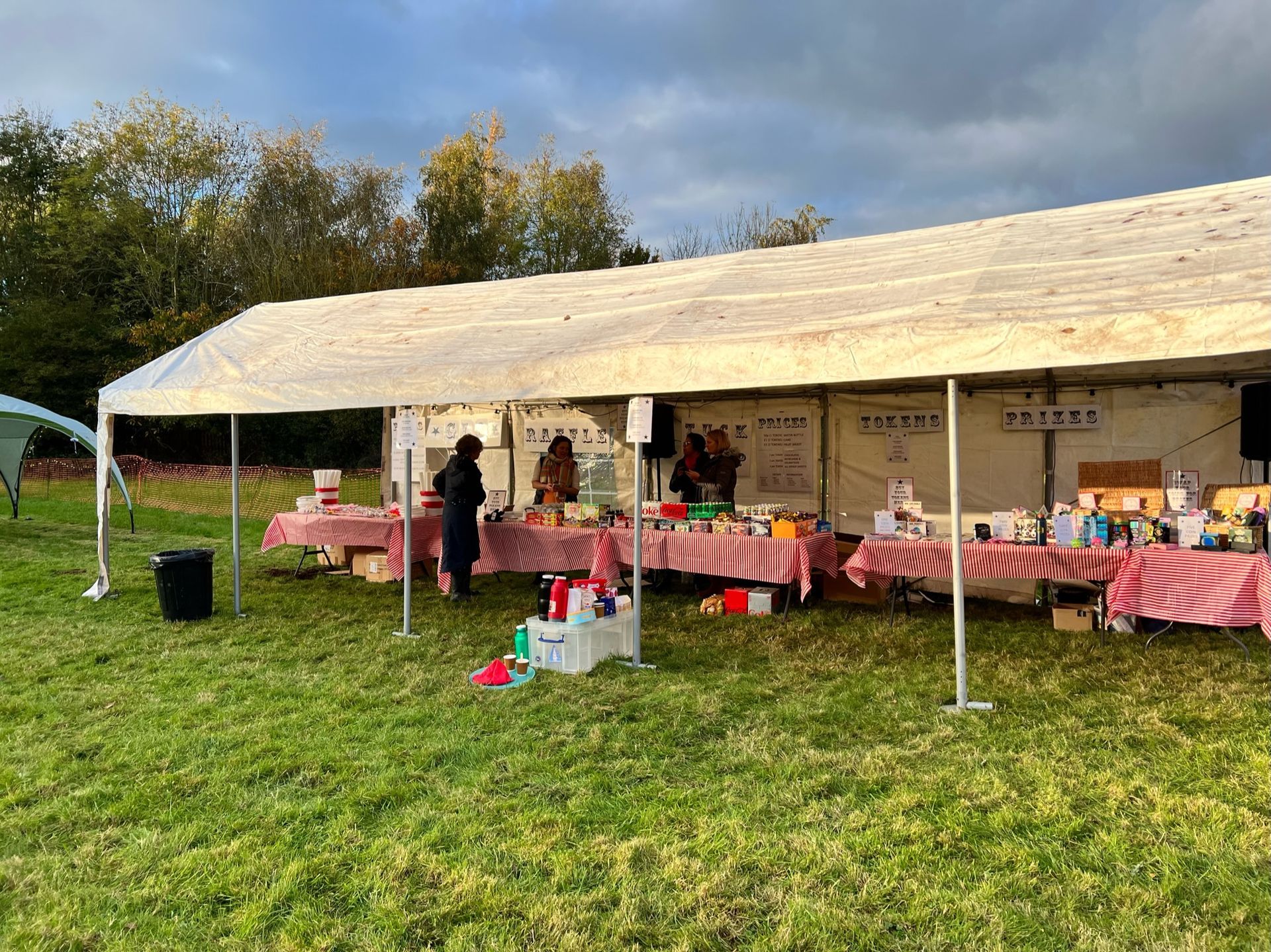 A white tent with vendors selling goods on red-checkered tables on a grassy field. Overcast sky.