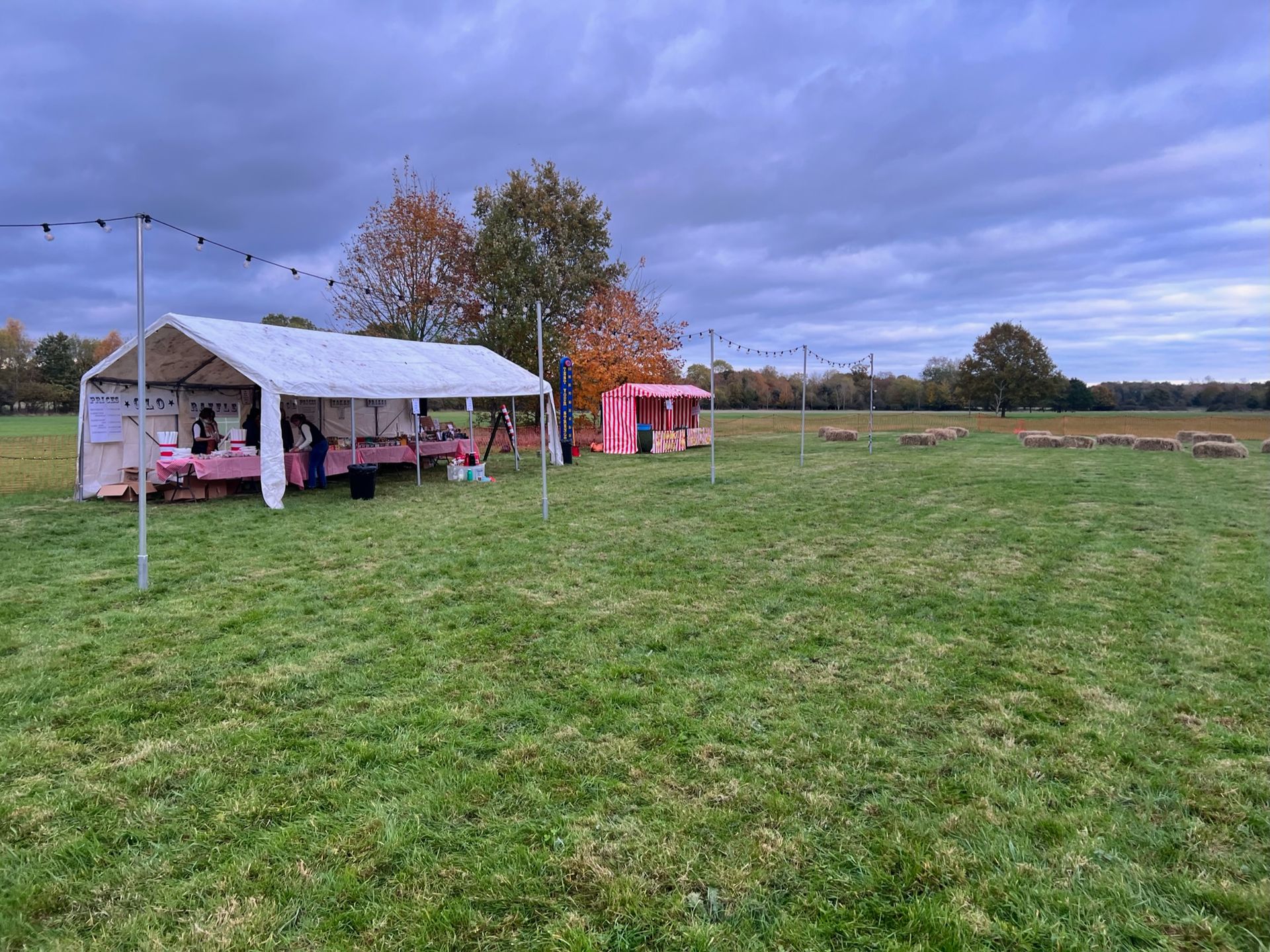 A white tent and a red food stall on a grassy field under a cloudy sky.