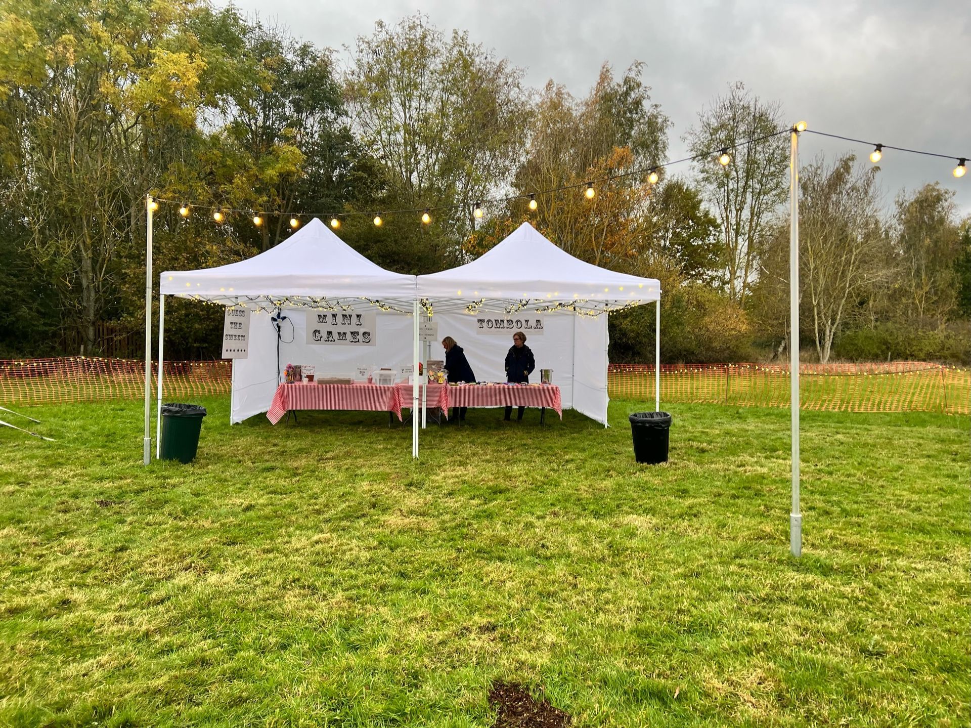 Two white tents set up in a grassy field with people behind a table, string lights, and fall foliage.