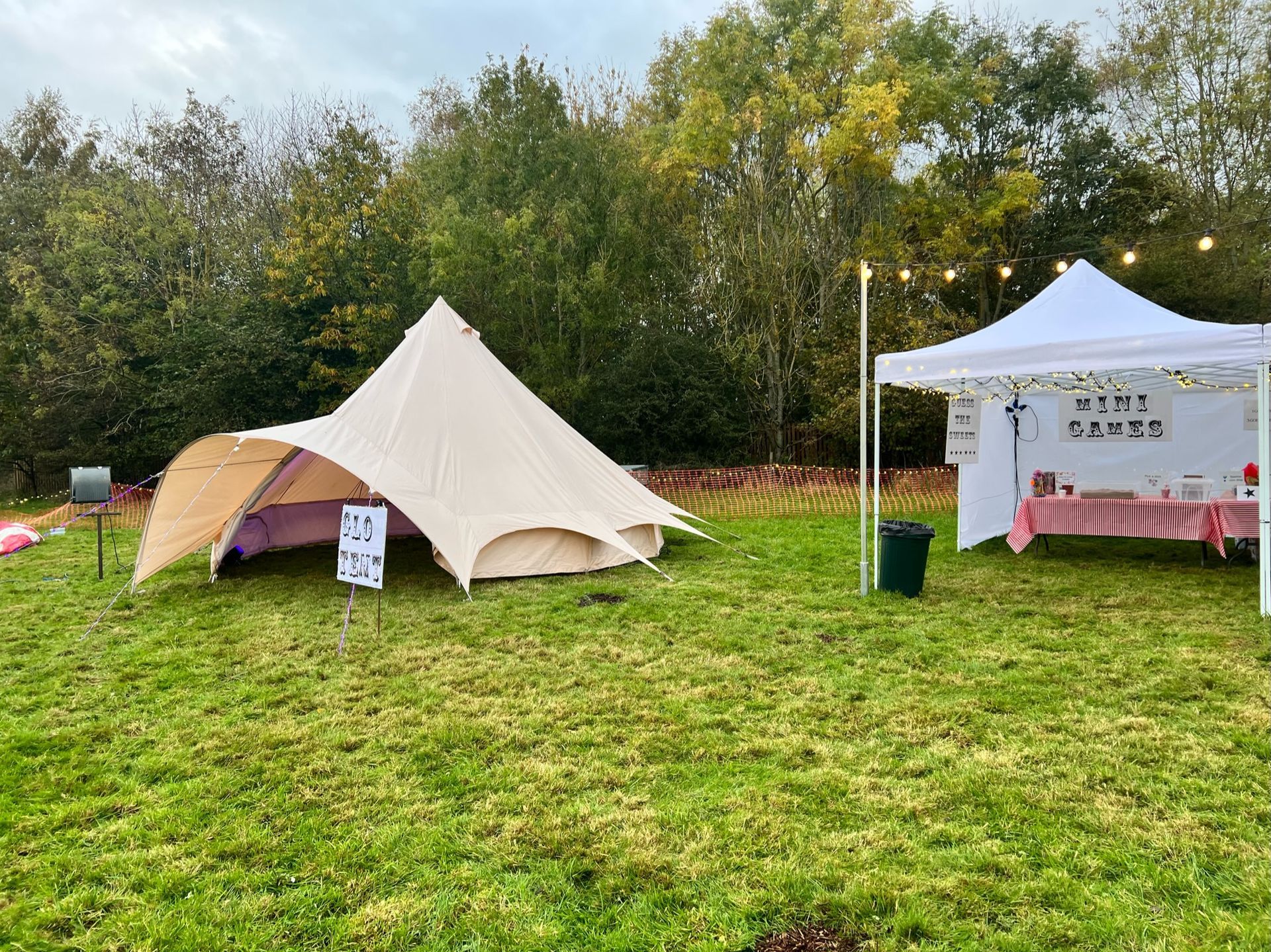 A beige bell tent and white market stall set up on a grassy field. String lights and trees in the background.