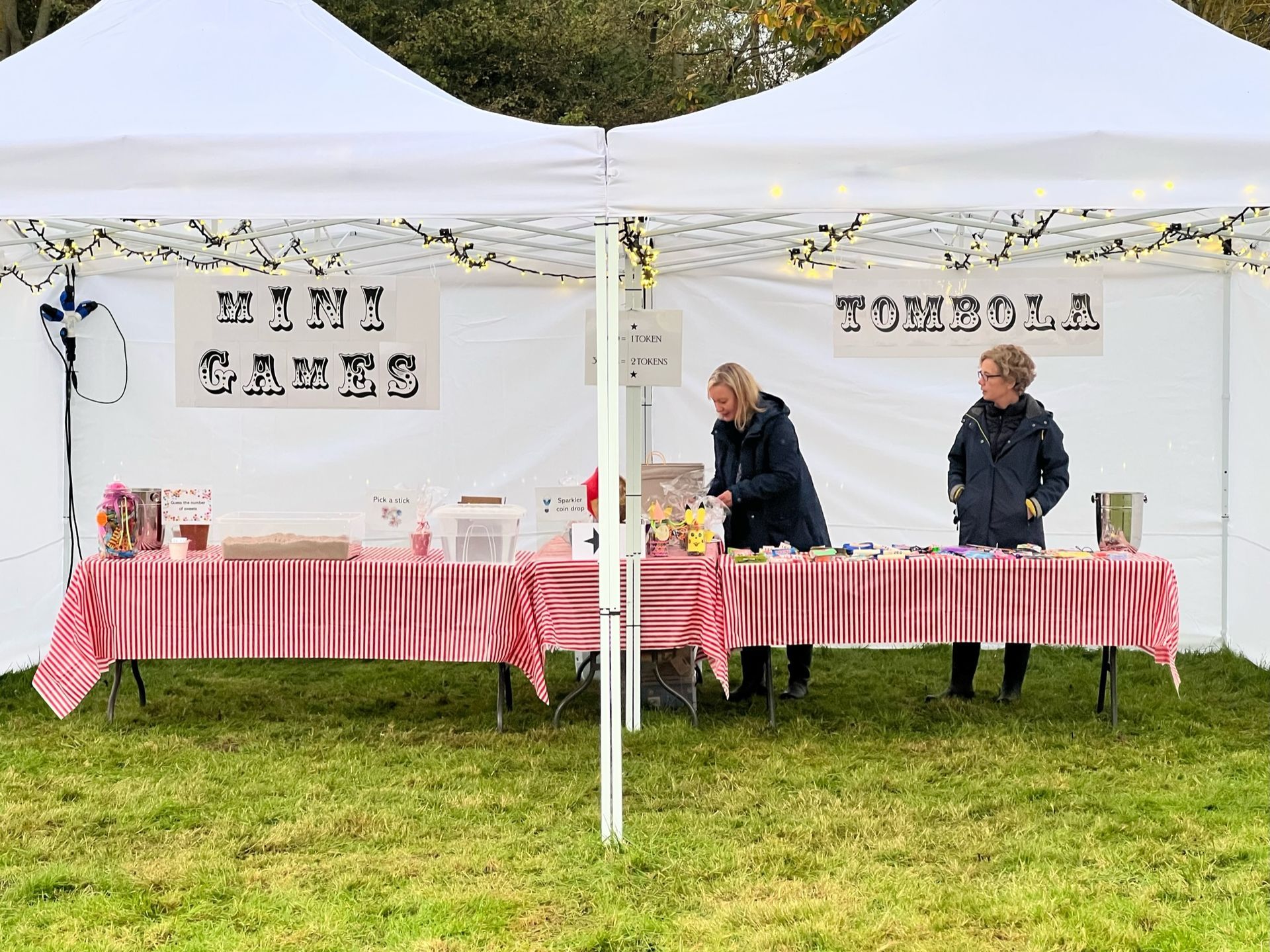 Two women at a Mini Games and Tombola stall under a white tent with colorful sweets.