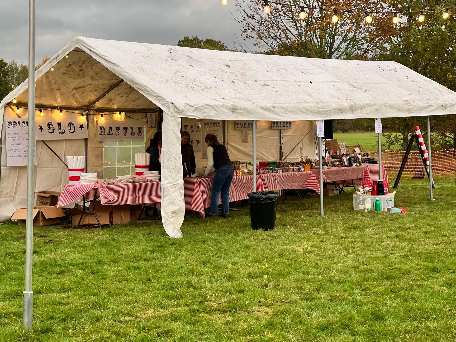 A tent with a food market set up outdoors. Two people stand behind tables covered in food and items.