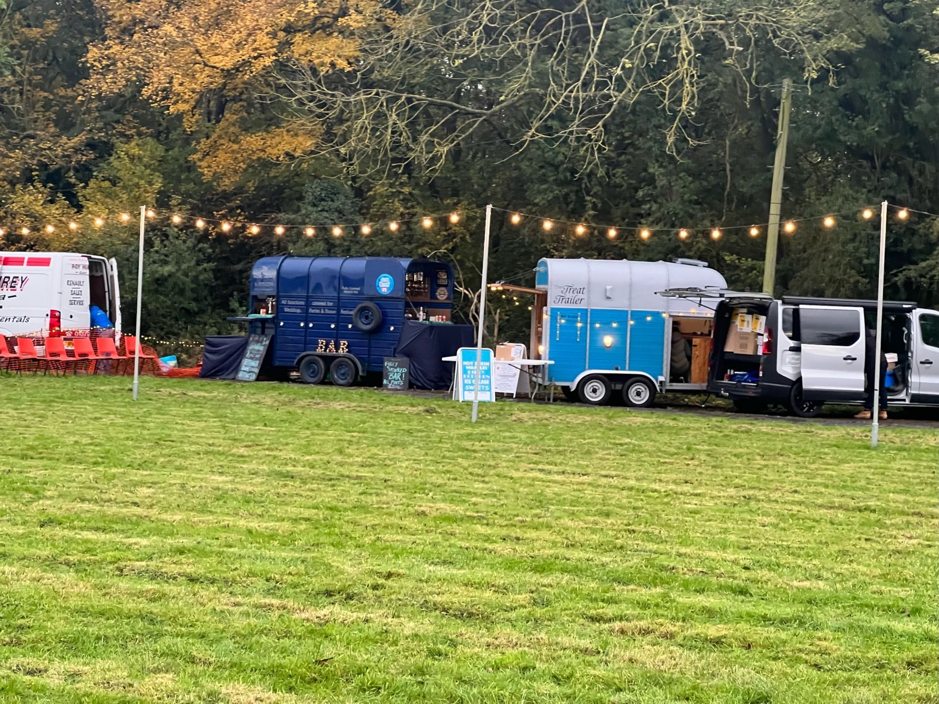 Food trucks lined up on a grassy field, strung with lights, selling food at an outdoor event.
