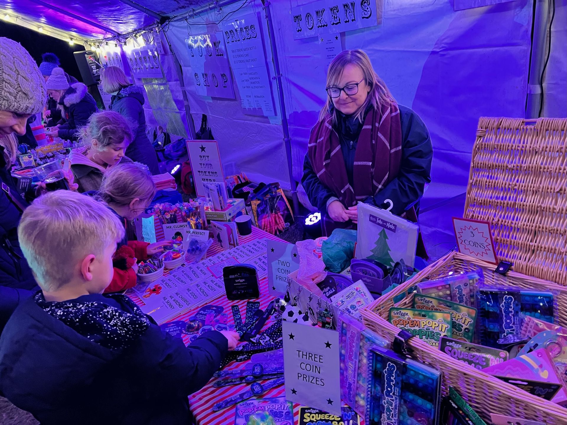 Woman at a craft market stall surrounded by customers, mostly children. Brightly lit with various crafts for sale.