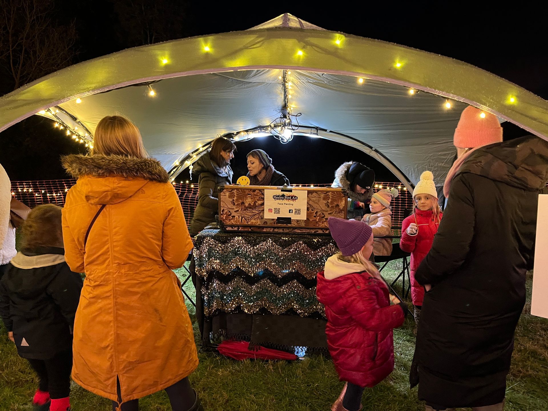 People at an outdoor illuminated market stall at night. Customers in warm clothes interacting with vendors.