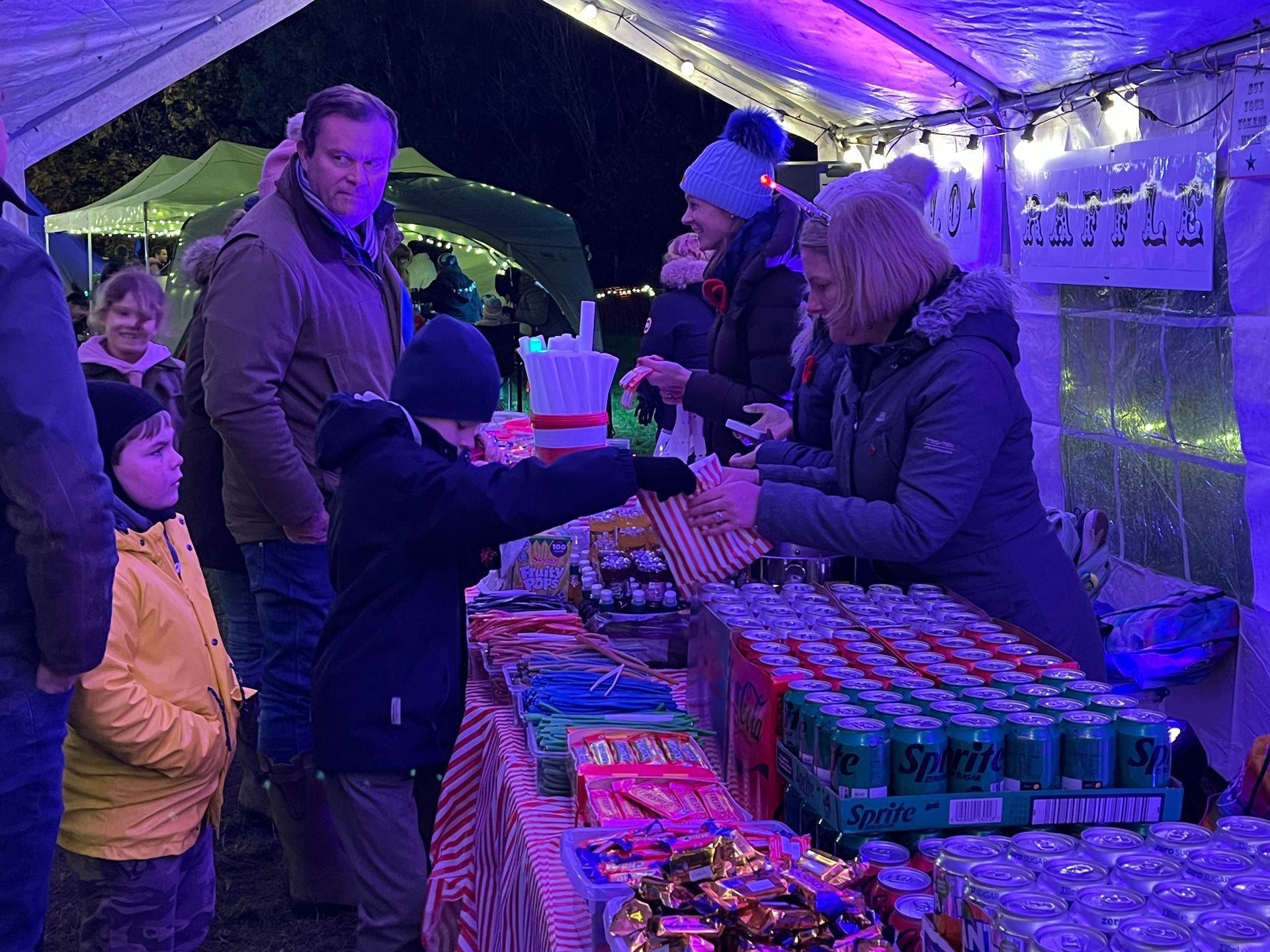 People buying snacks and drinks at a festive outdoor stall at night, including a child reaching toward the vendor.