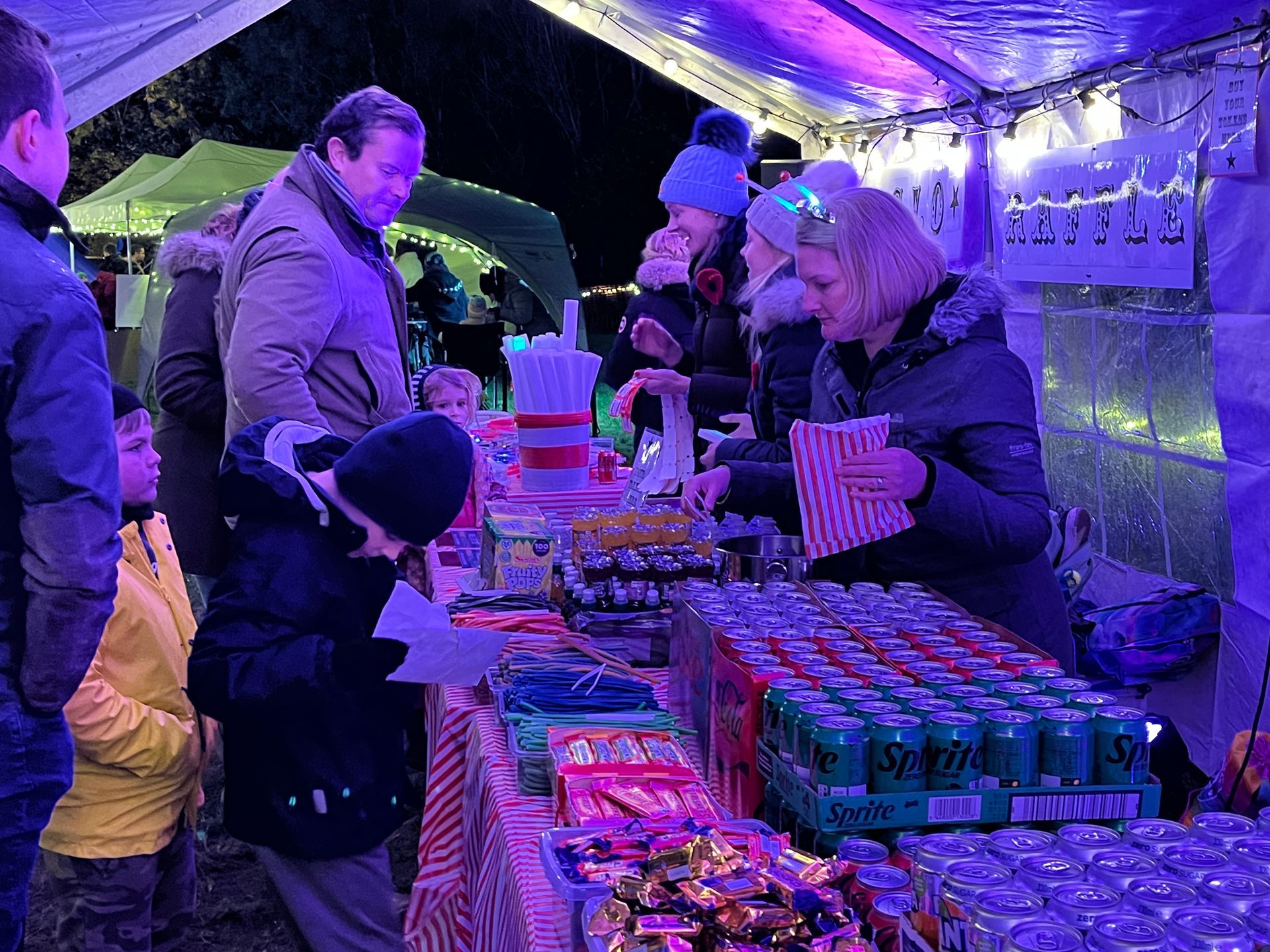 People buying snacks at a brightly lit outdoor food stall, at night.