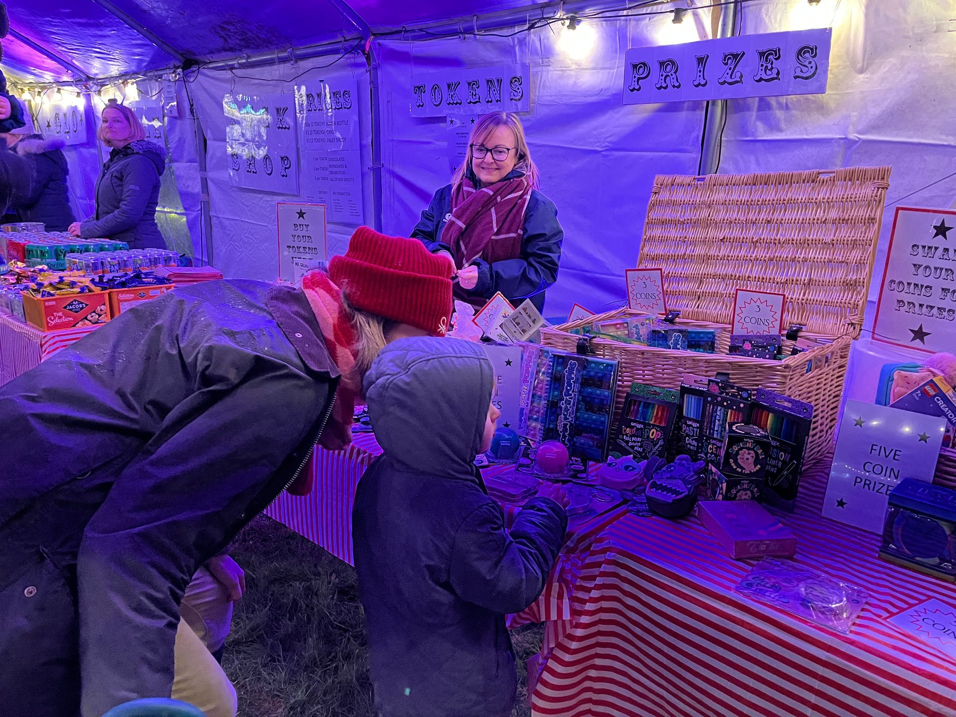 A person and child looking at crafts at a market stall, woman behind the counter, lit by purple lights.