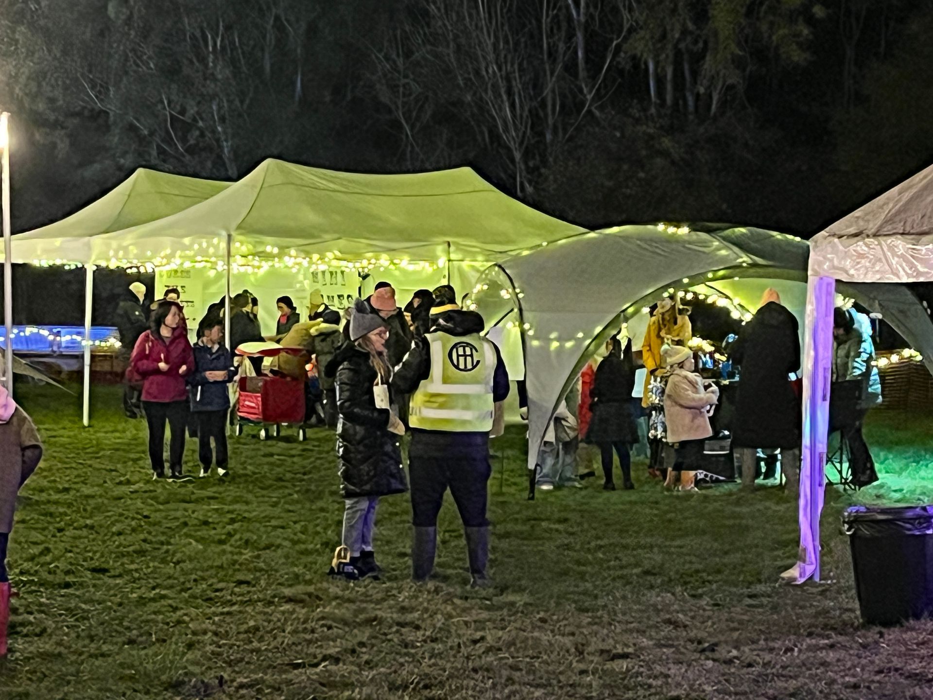 Nighttime outdoor event with tents, people, and string lights on a grassy field.