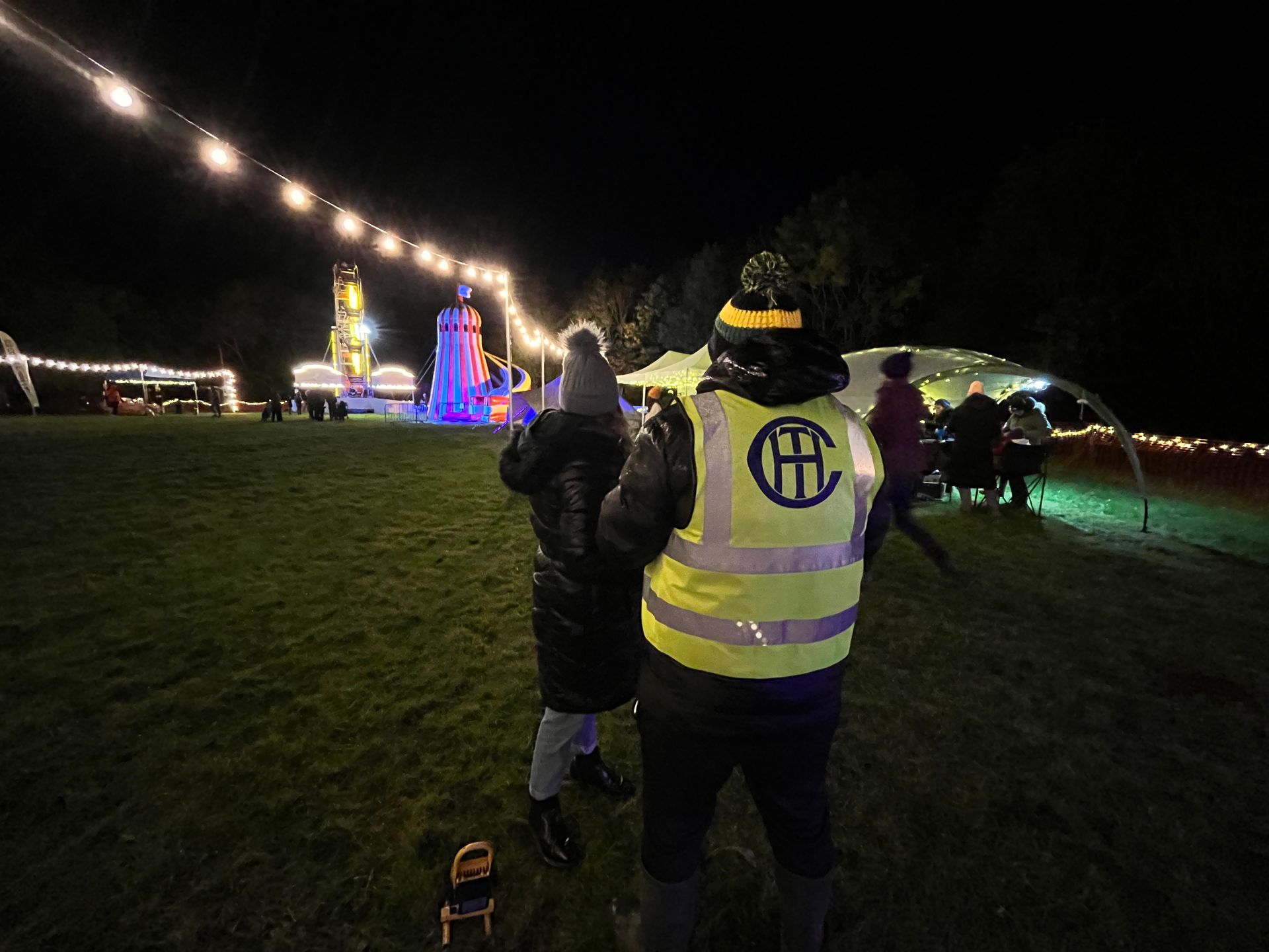 People at a nighttime outdoor event. A person in a reflective vest and hat. Lights and a structure are in the background.