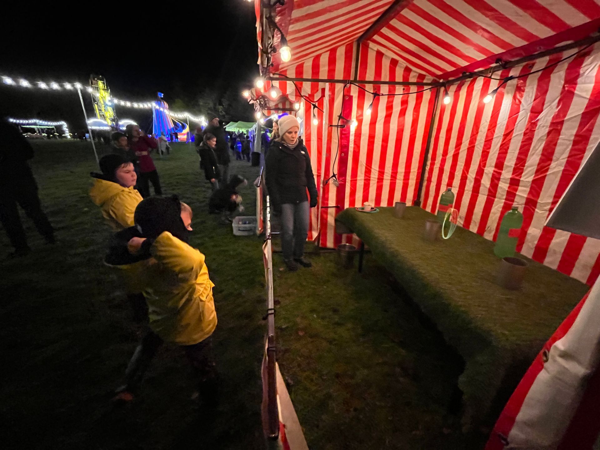 People playing a carnival game at night, with red and white striped tent and string lights.