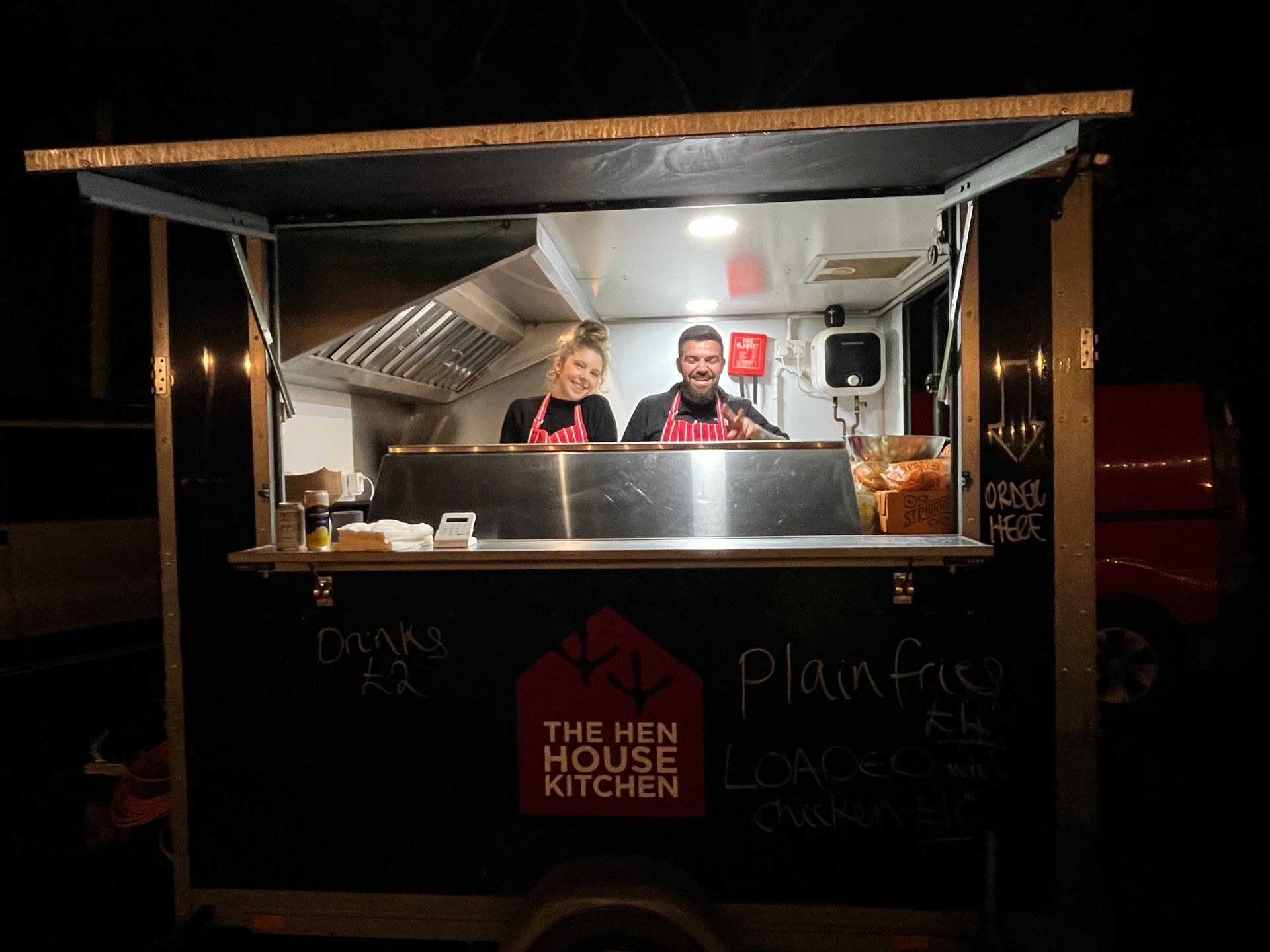 Two people in a food truck: a woman and a man smiling. Truck has a black exterior, illuminated interior.