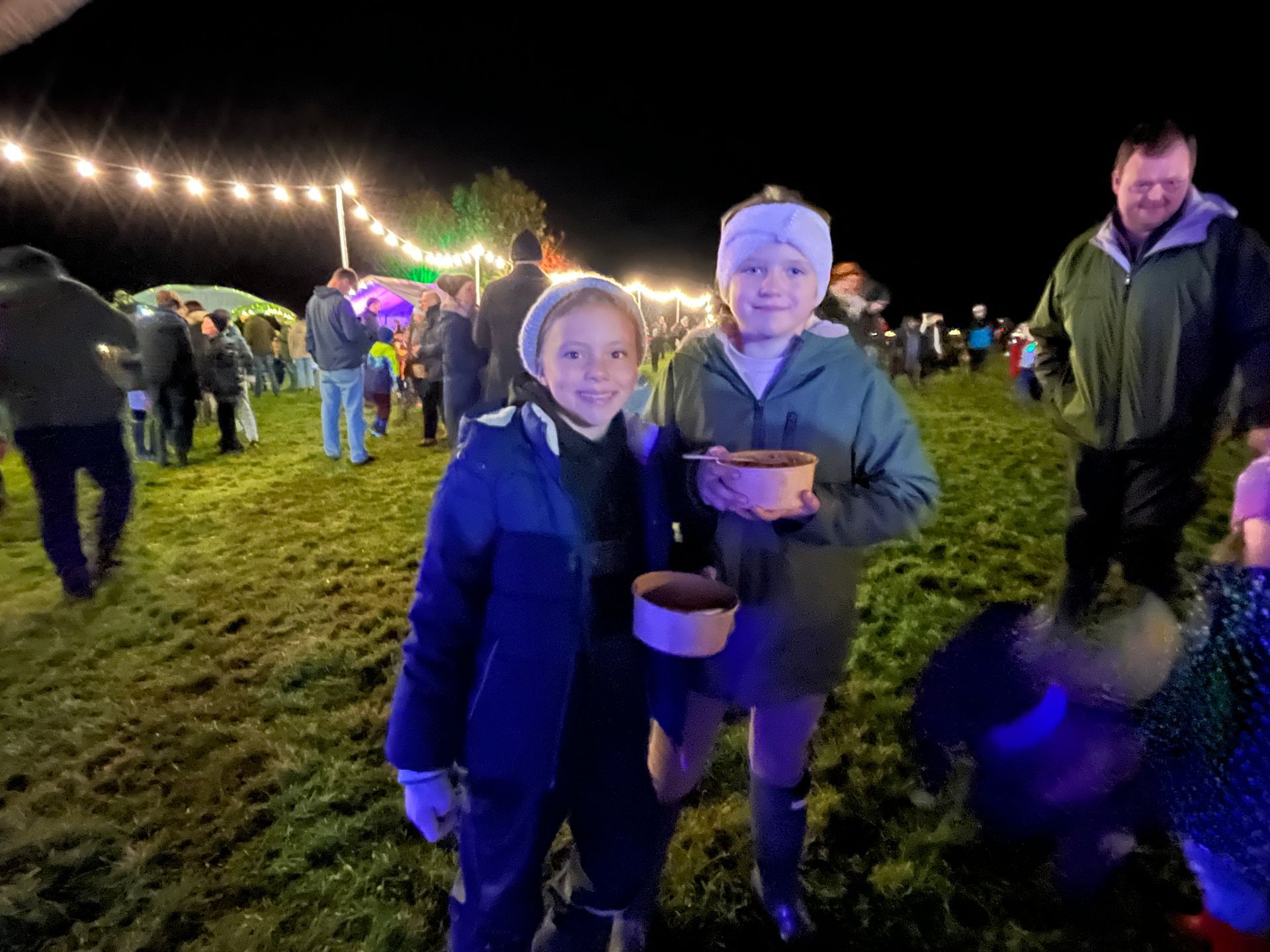 Two children at night event, holding food. Crowd and lights in background.