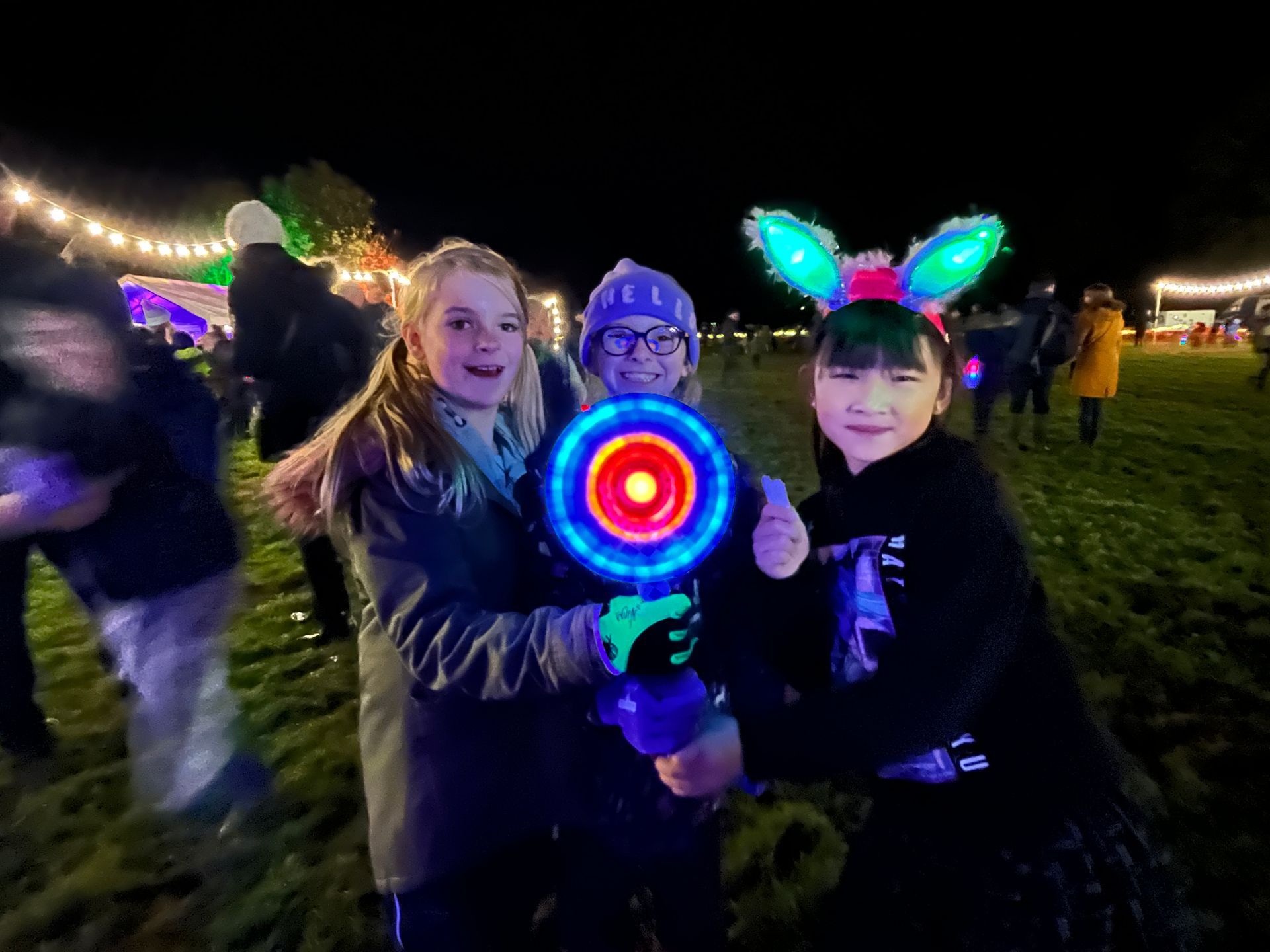 Three smiling kids holding a glowing toy, outdoors at night; festive lights, grass field.