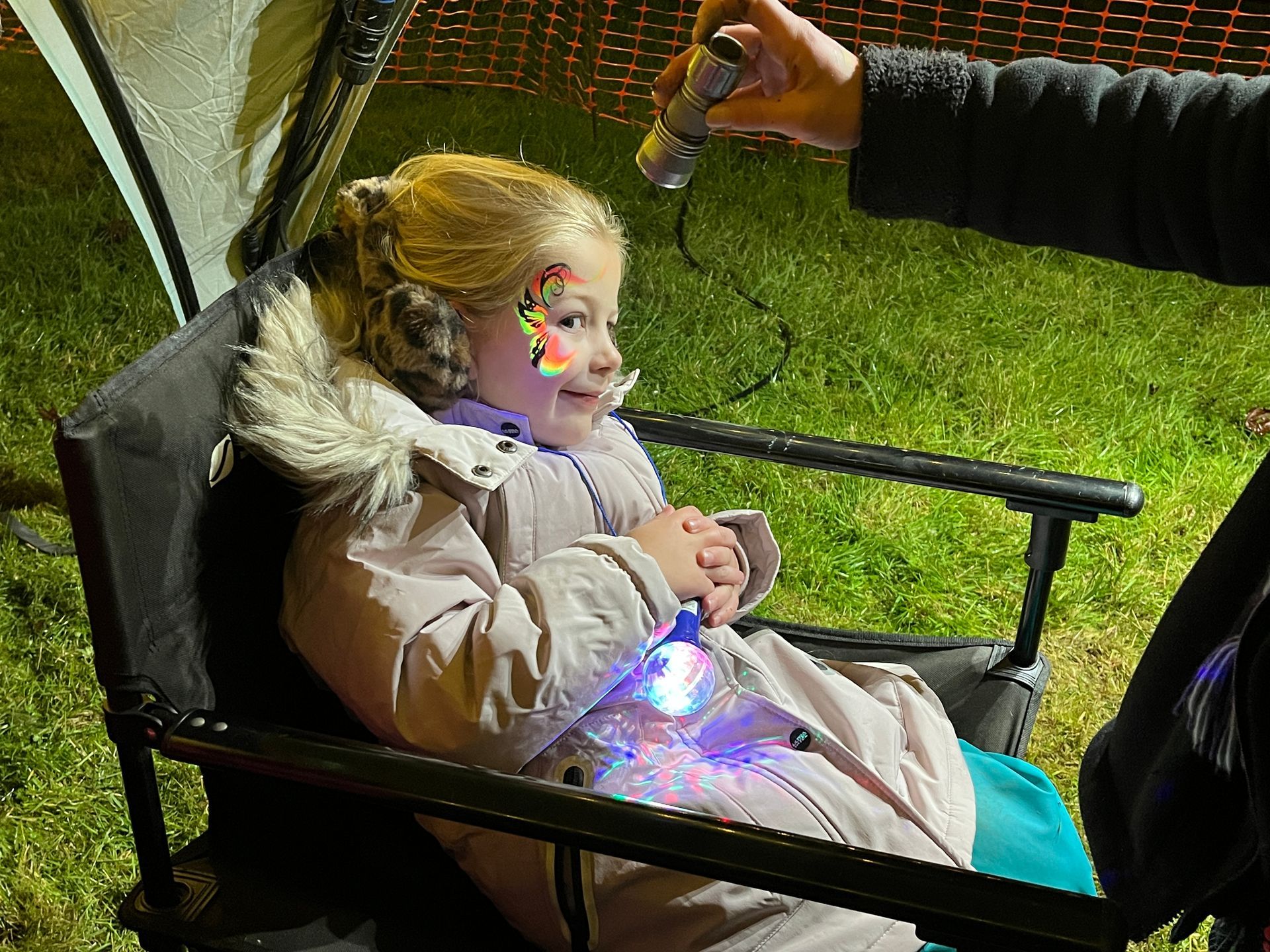 Girl with face paint sits in chair, someone adds a light to her arm. Outdoors at night.