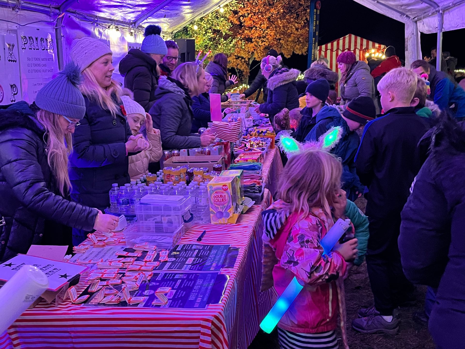 People browse candy at a nighttime market stall. Children wear light-up toys. Colorful treats are displayed.