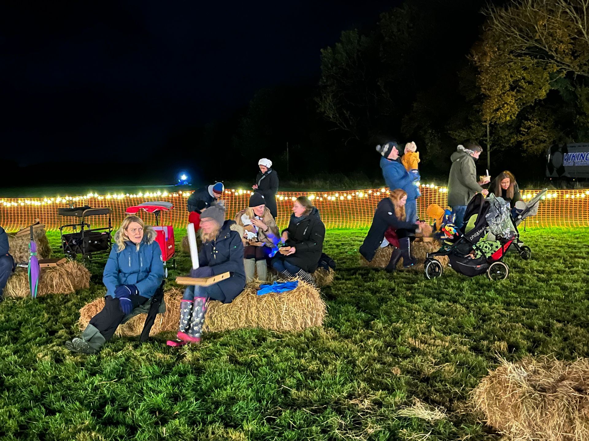 People sitting on hay bales outdoors at night. Festive lights decorate the field.