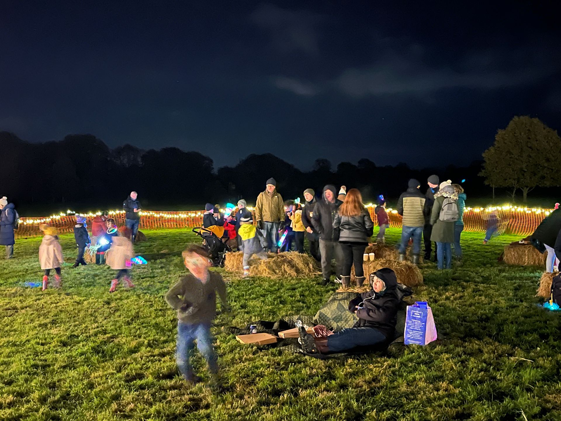 People gather at night in a field, lit by string lights. Some sit on hay bales, others stand.