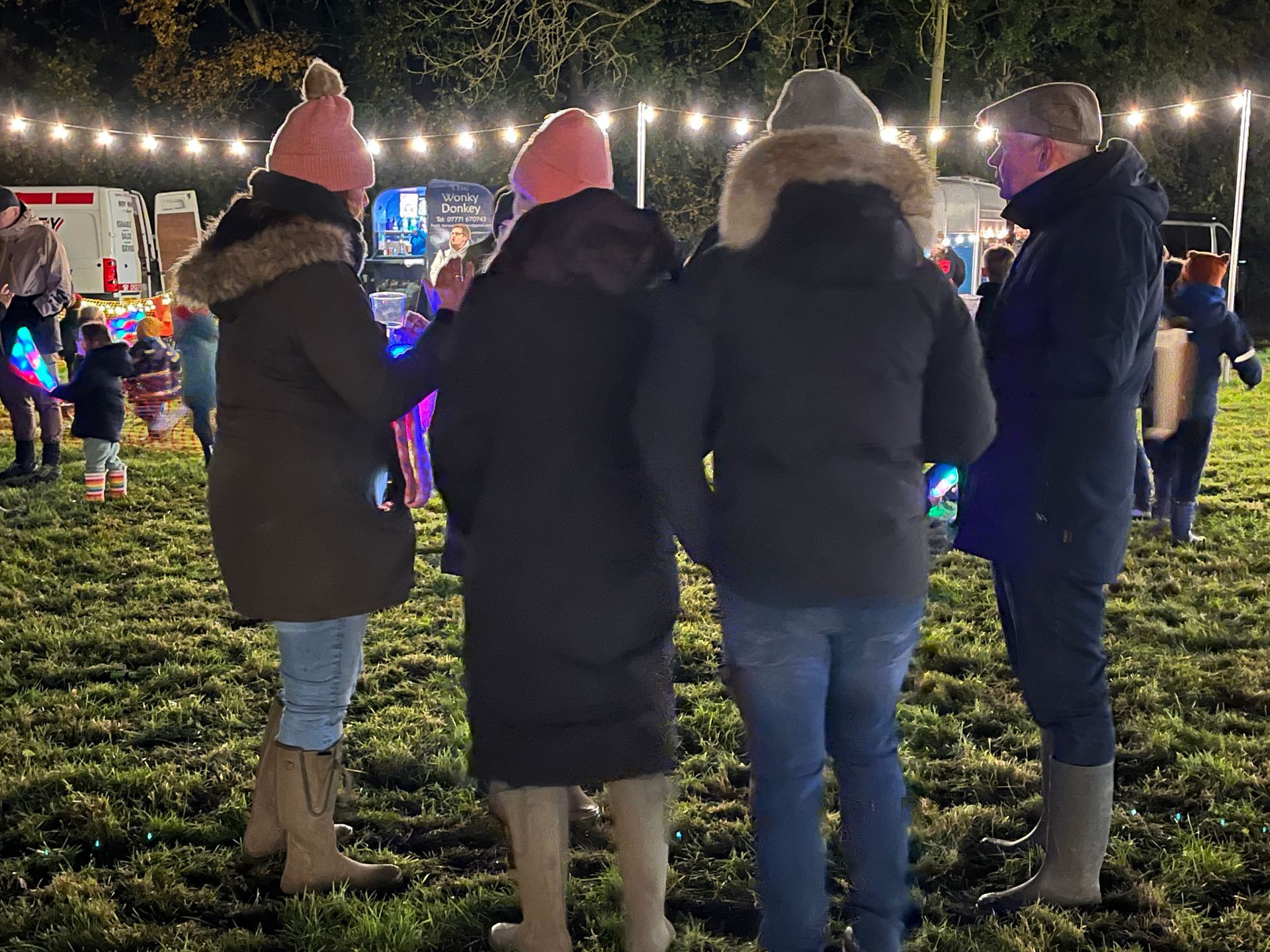 People in coats and hats stand outdoors at a night event with lights strung above.