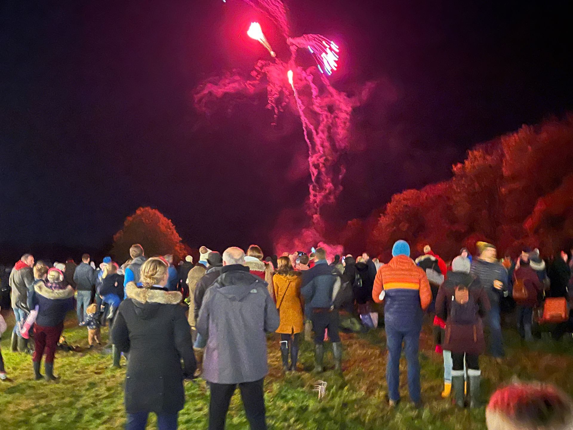 People watching fireworks in a field at night, with red explosions and smoke.