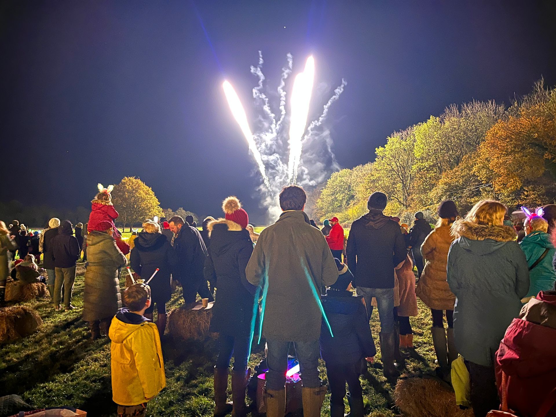 People watching fireworks at night in a field. Some children on shoulders, fall foliage visible.