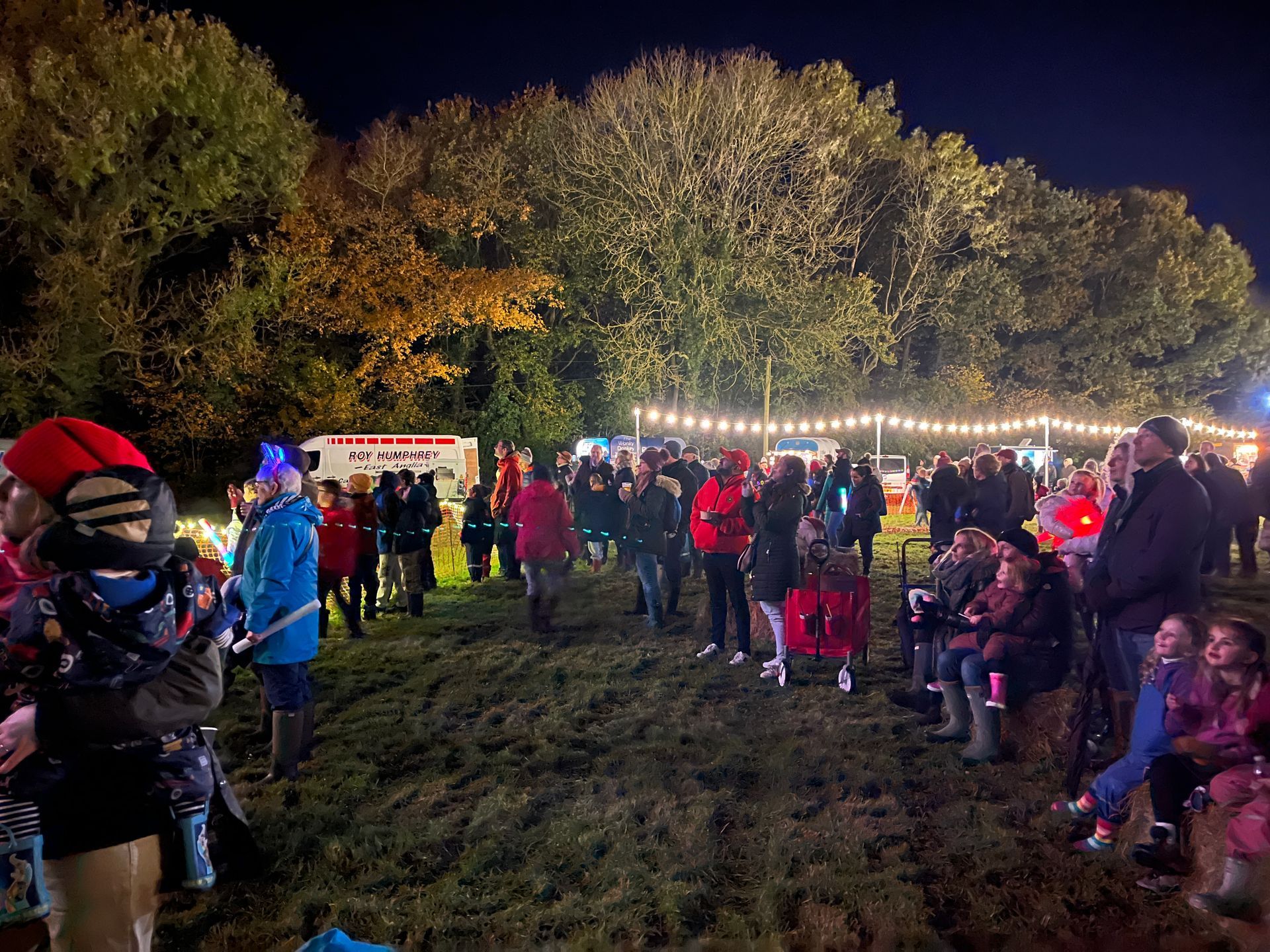Nighttime outdoor gathering with crowd, food stall, and strung lights. Trees in the background.