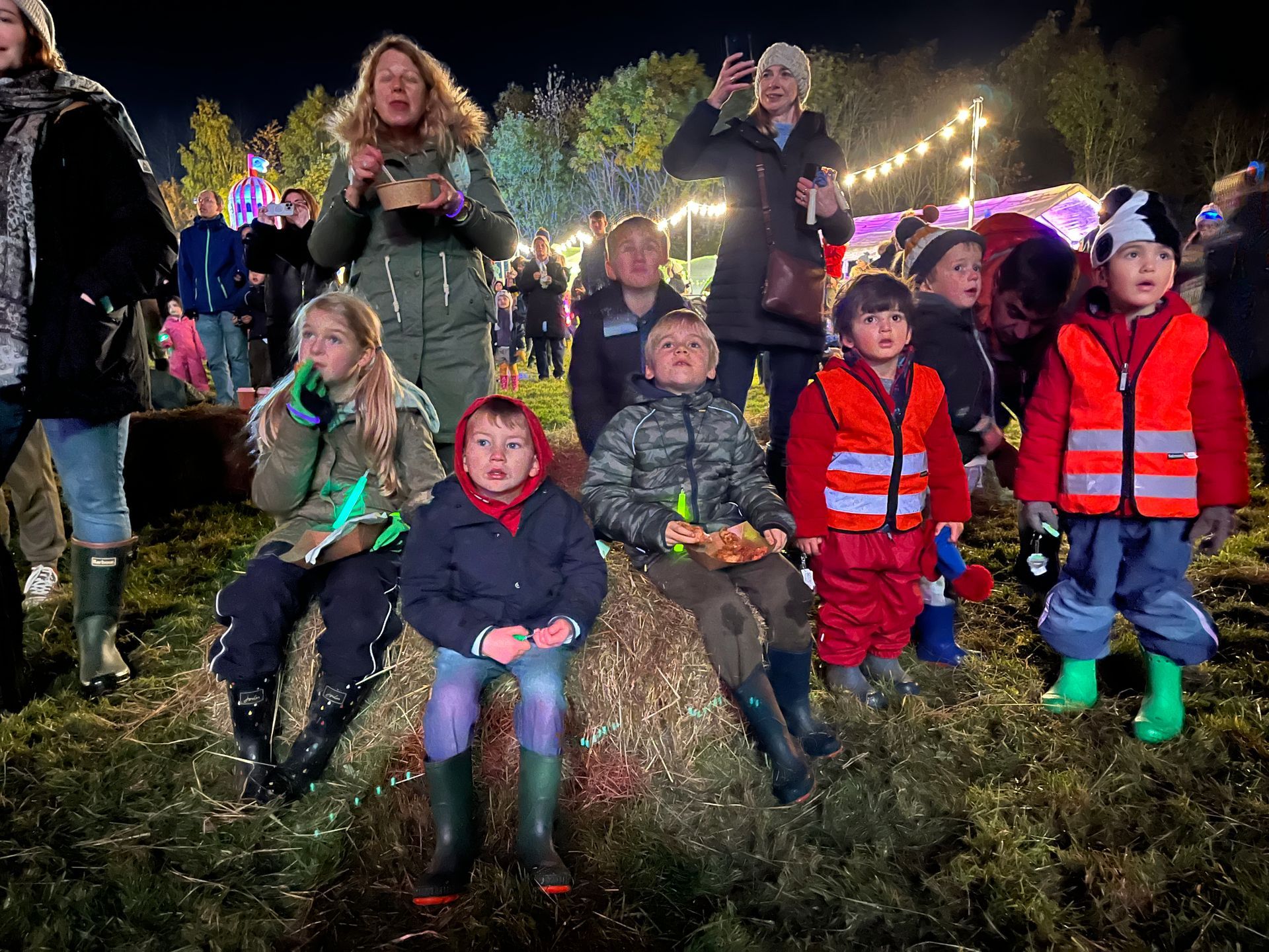 Children sit on hay bales at an outdoor night event. People watch in background.