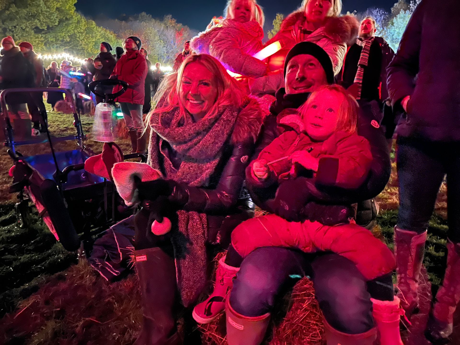 Family at night, smiling. Mother and child seated, father holds child. Outdoor festival, lights, hay bales.