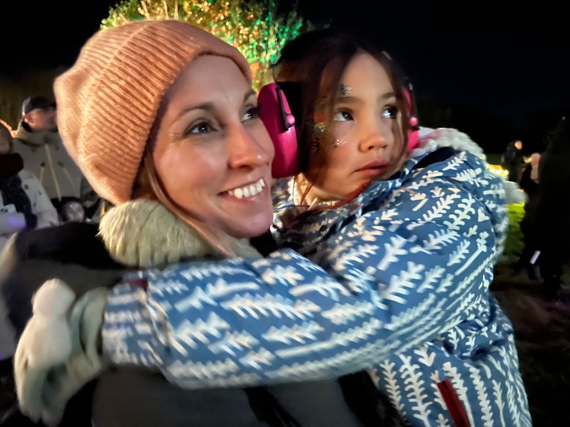 Woman hugs a child, both smiling at a nighttime event. The child wears earmuffs and a winter coat.