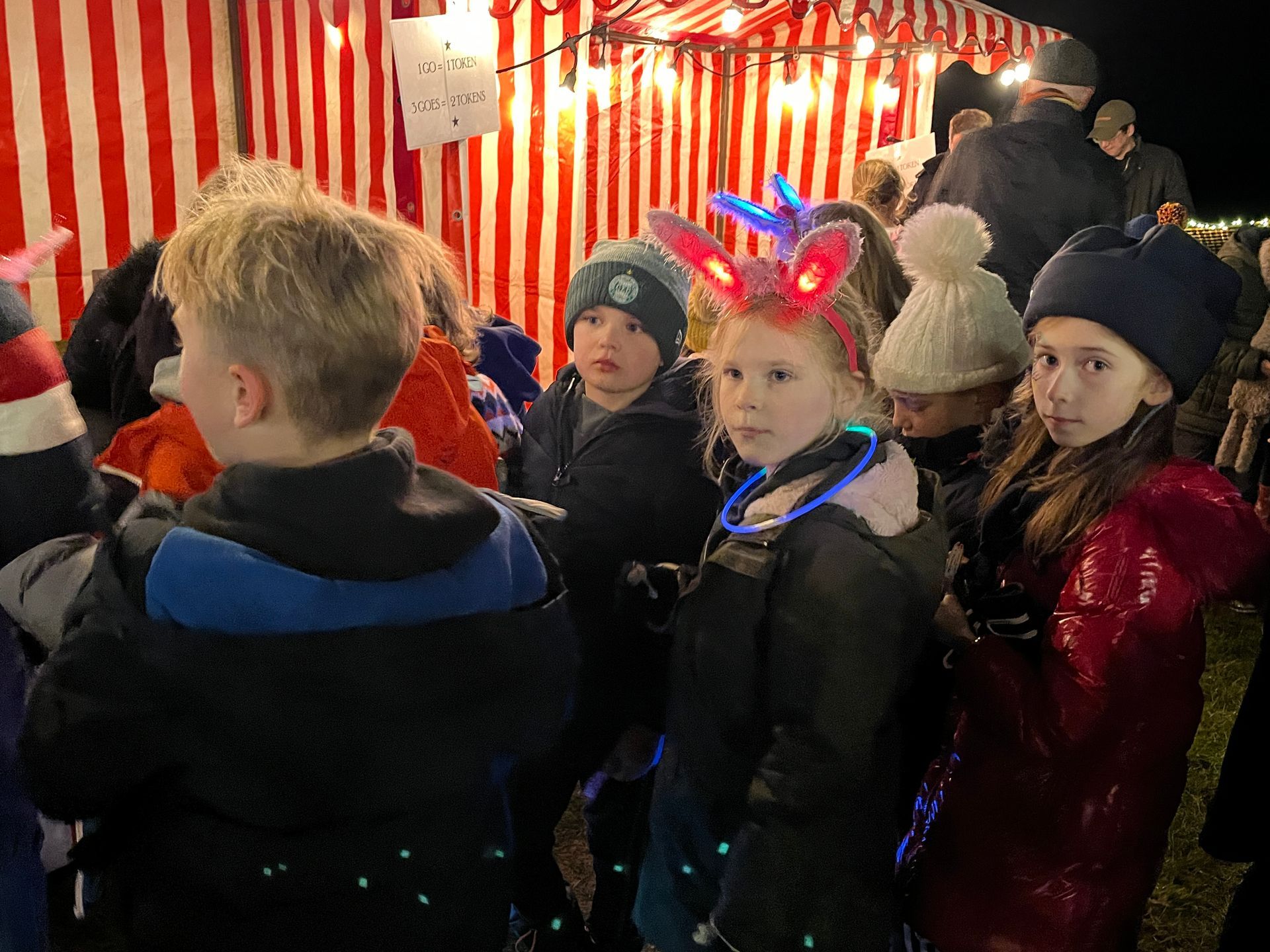 Children in winter coats and hats, some with light-up accessories, stand near a striped tent at night.