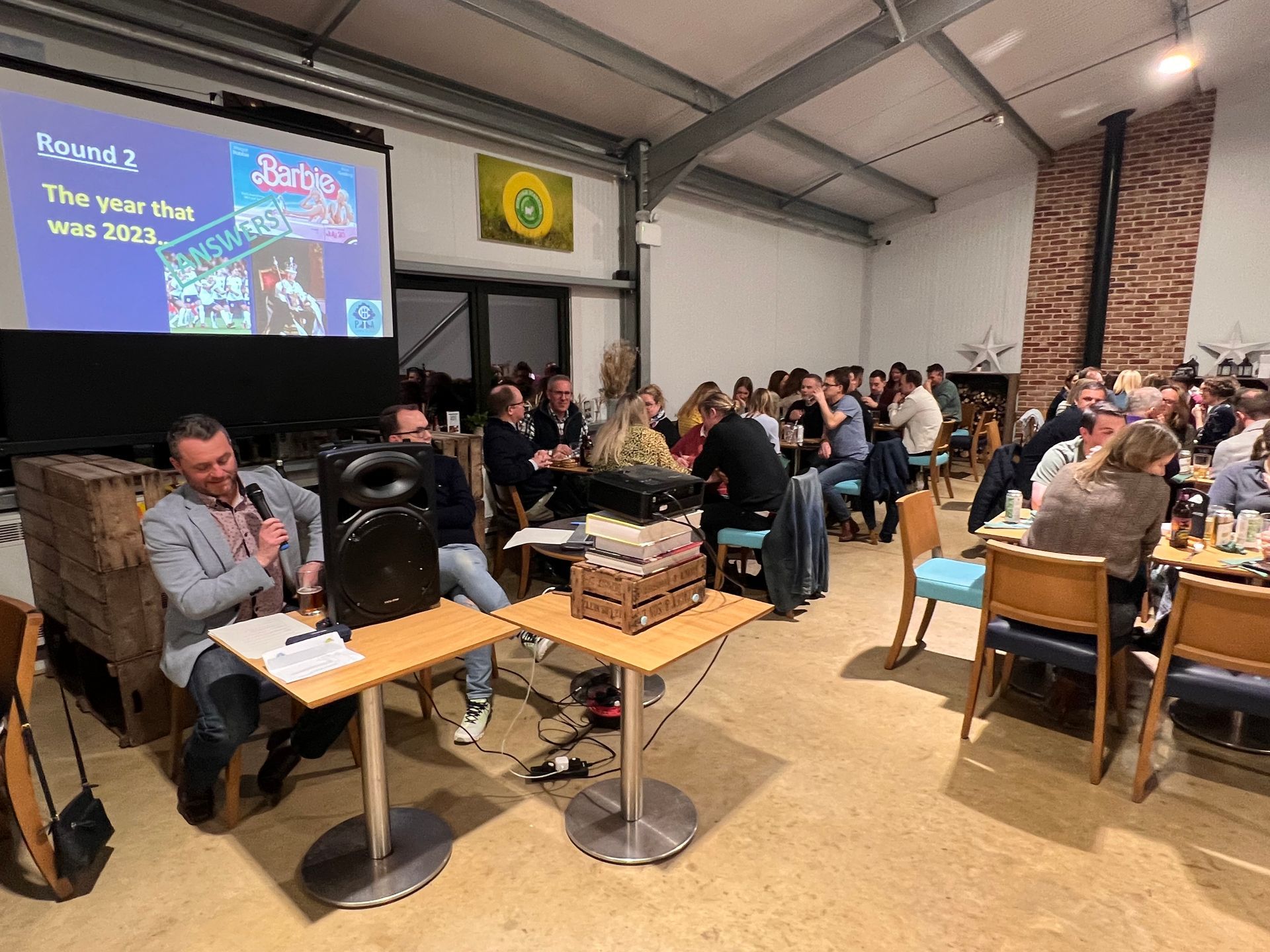 A group of people at a trivia night in a well-lit room. A man speaks into a microphone while others watch.